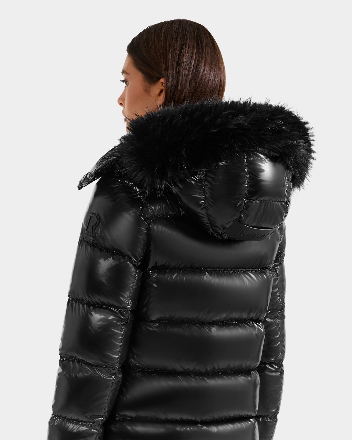 A person with brown hair, seen from behind, wears the Rudsak BREE S - L125583 Black women’s down puffer jacket with a shiny, weather-resistant finish and black faux fur hood, against a plain light gray background.