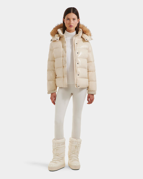 A woman faces forward in the Rudsak BREE S - L125583 Vanilla women’s down puffer with ultra-gloss finish and a removable fur-trimmed hood, styled with a cream turtleneck, fitted pants, and tall padded snow boots against a white background.