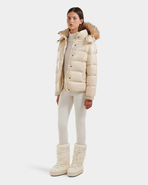 A woman faces forward in the Rudsak BREE S - L125583 Vanilla women’s down puffer with an ultra-gloss finish, fur-trimmed removable hood, matching fitted pants, and large cream snow boots against a plain white background.