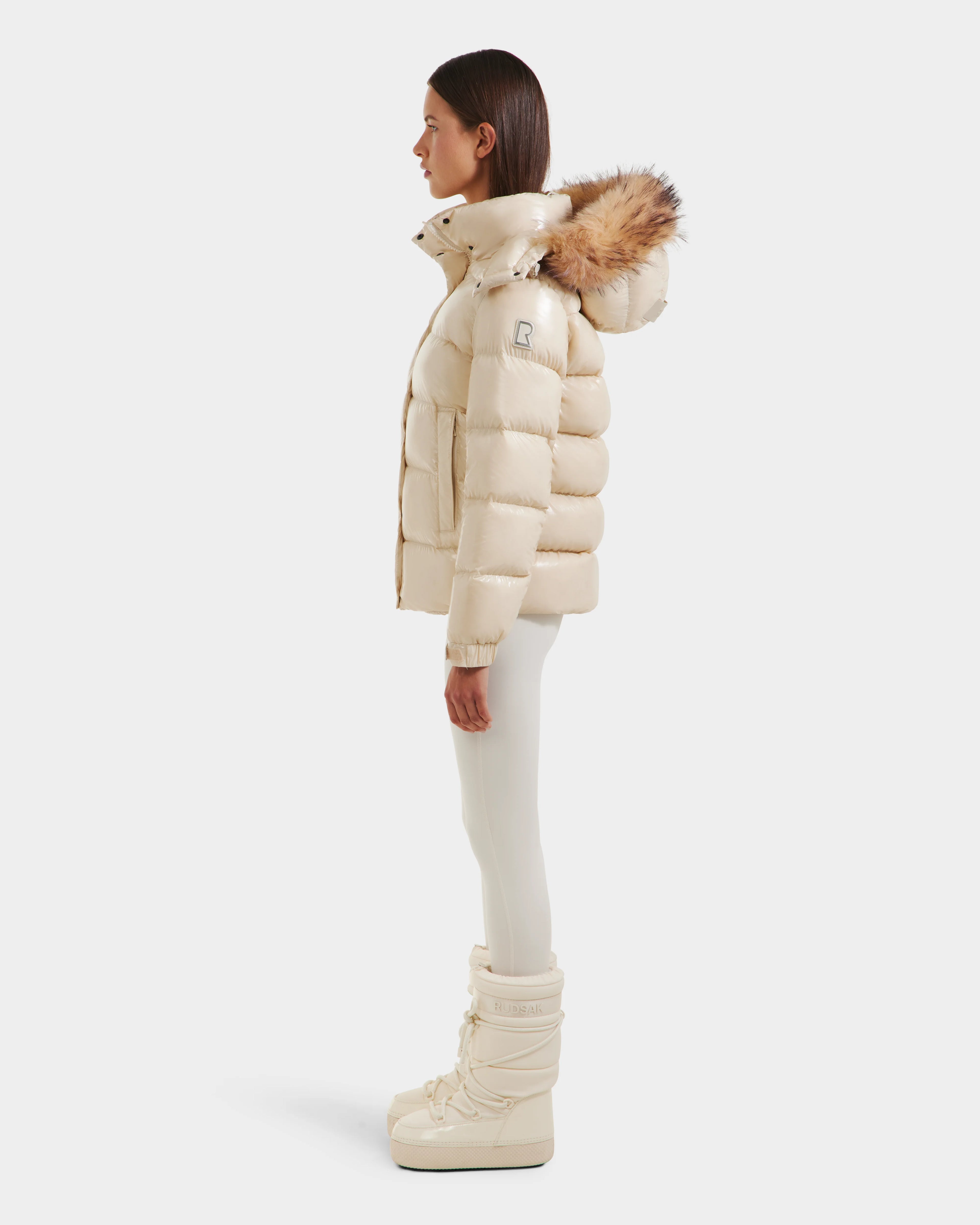 A woman stands in profile wearing the Rudsak BREE S - L125583 Vanilla women’s down puffer jacket with a removable fur-trimmed hood, matching leggings, and winter boots against a plain white background.