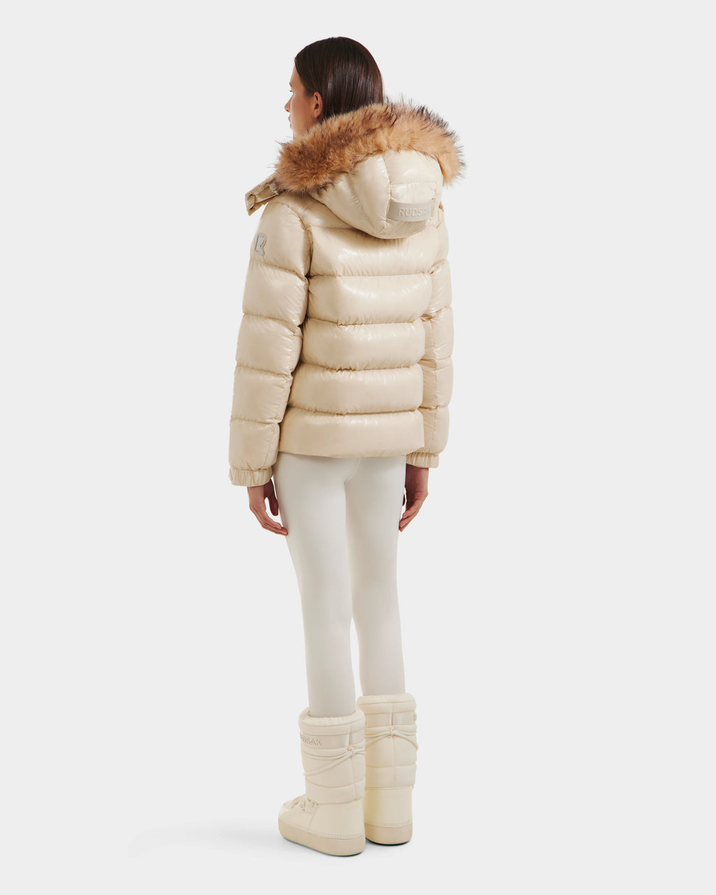 A woman, seen from behind, wears the Rudsak BREE S - L125583 Vanilla women’s down puffer jacket with a shiny cream finish and removable fur-trimmed hood, paired with cream leggings and snow boots against a plain white background.