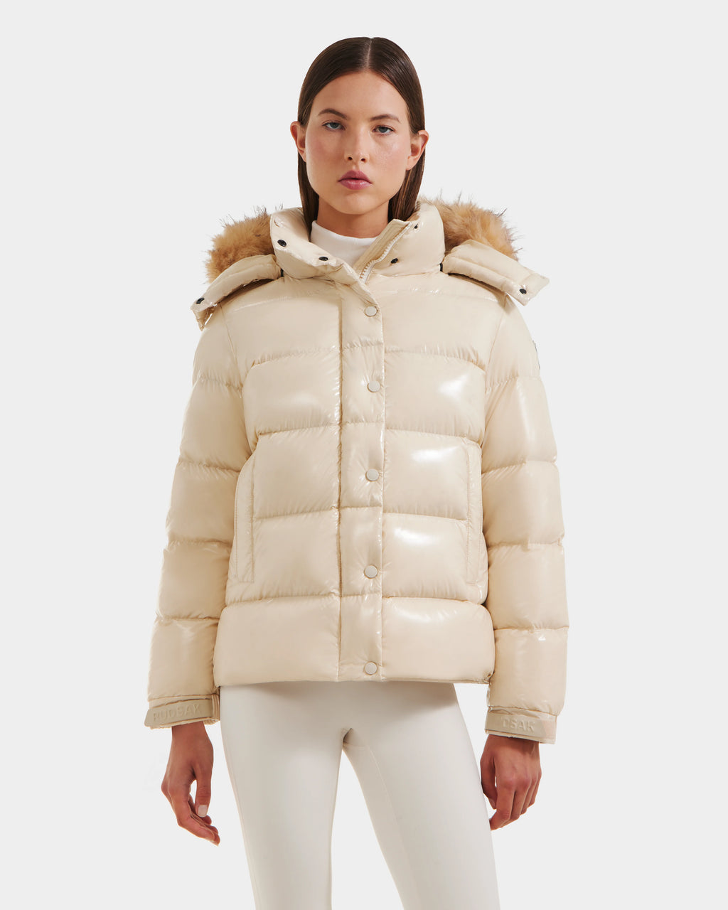 A woman stands facing forward against a white background, wearing the Rudsak BREE S - L125583 Vanilla down puffer jacket with an ultra-gloss finish and faux fur-trimmed removable hood, styled with a white turtleneck and pants.