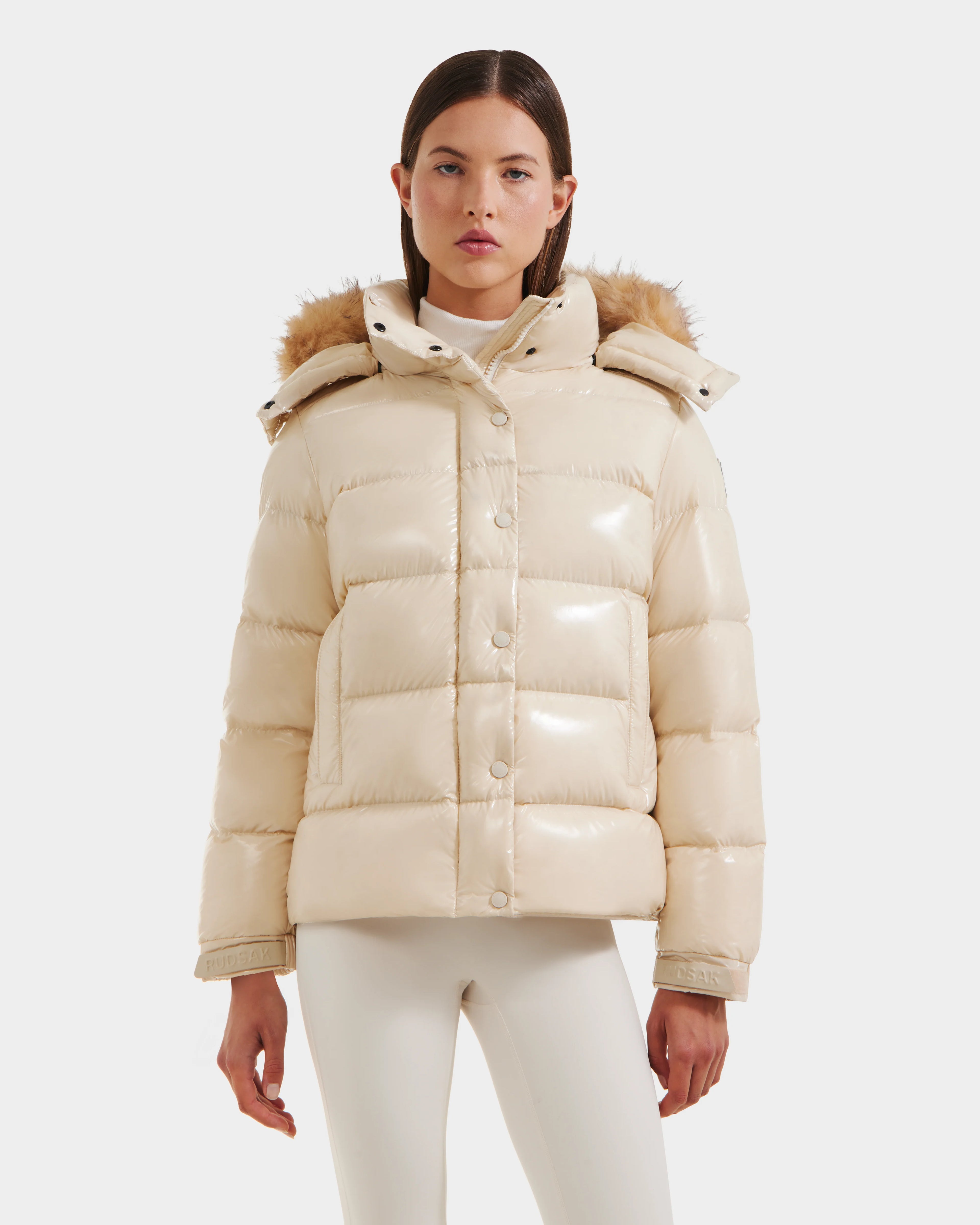 A woman stands facing forward against a white background, wearing the Rudsak BREE S - L125583 Vanilla down puffer jacket with an ultra-gloss finish and faux fur-trimmed removable hood, styled with a white turtleneck and pants.
