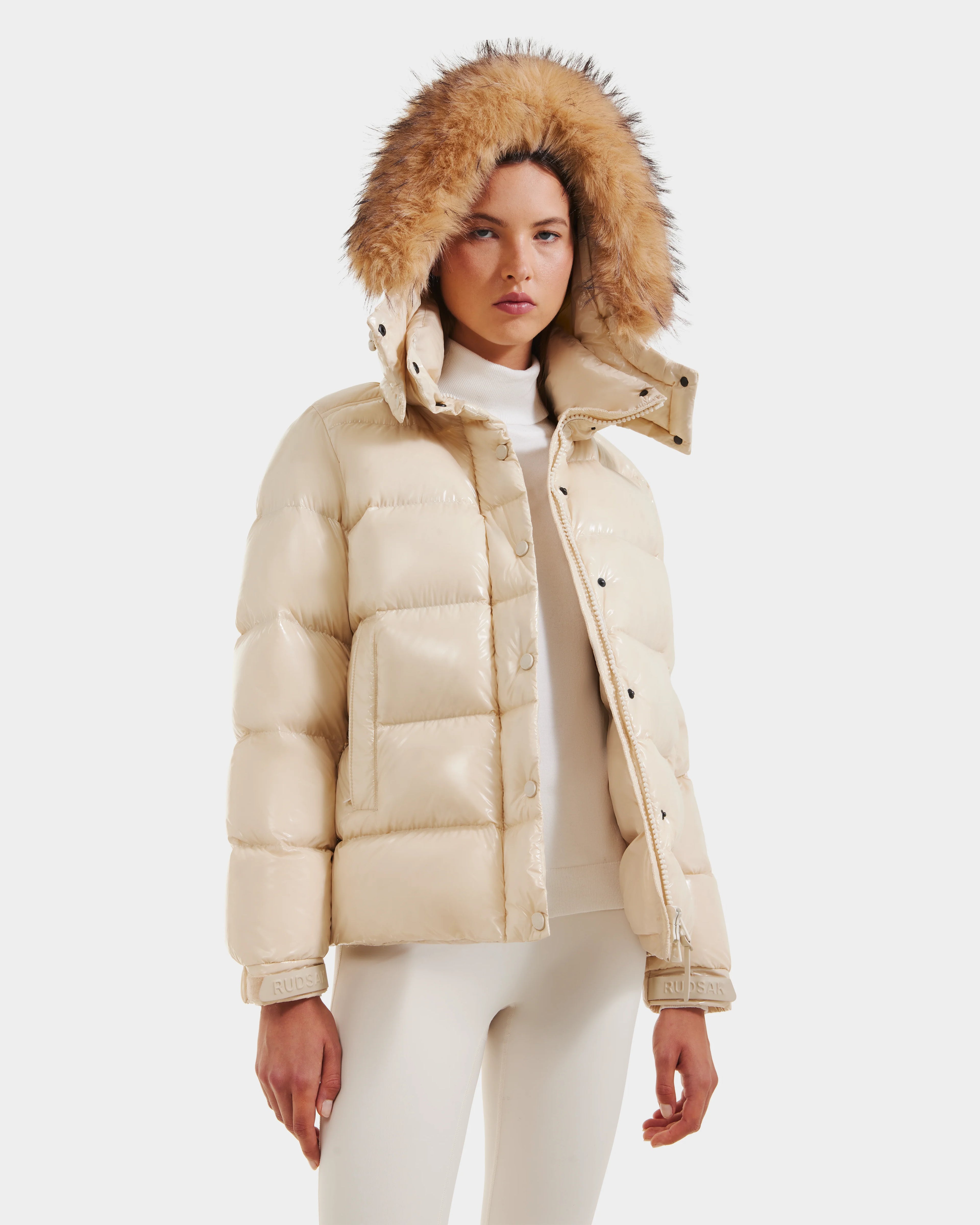 A woman wears the Rudsak BREE S - L125583 Vanilla down puffer with ultra-gloss finish and removable fur-lined hood, paired with a white turtleneck and cream pants, standing against a plain white background.
