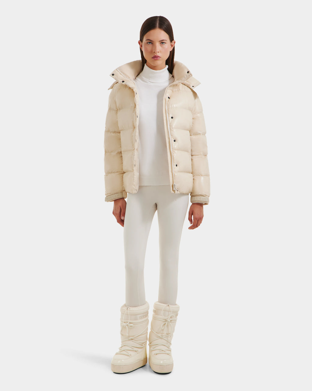 A woman with straight brown hair and a neutral expression stands facing forward against a white background, wearing the Rudsak BREE S - L125583 Vanilla women's down puffer with removable hood, cream snow boots, white turtleneck, and fitted white pants.