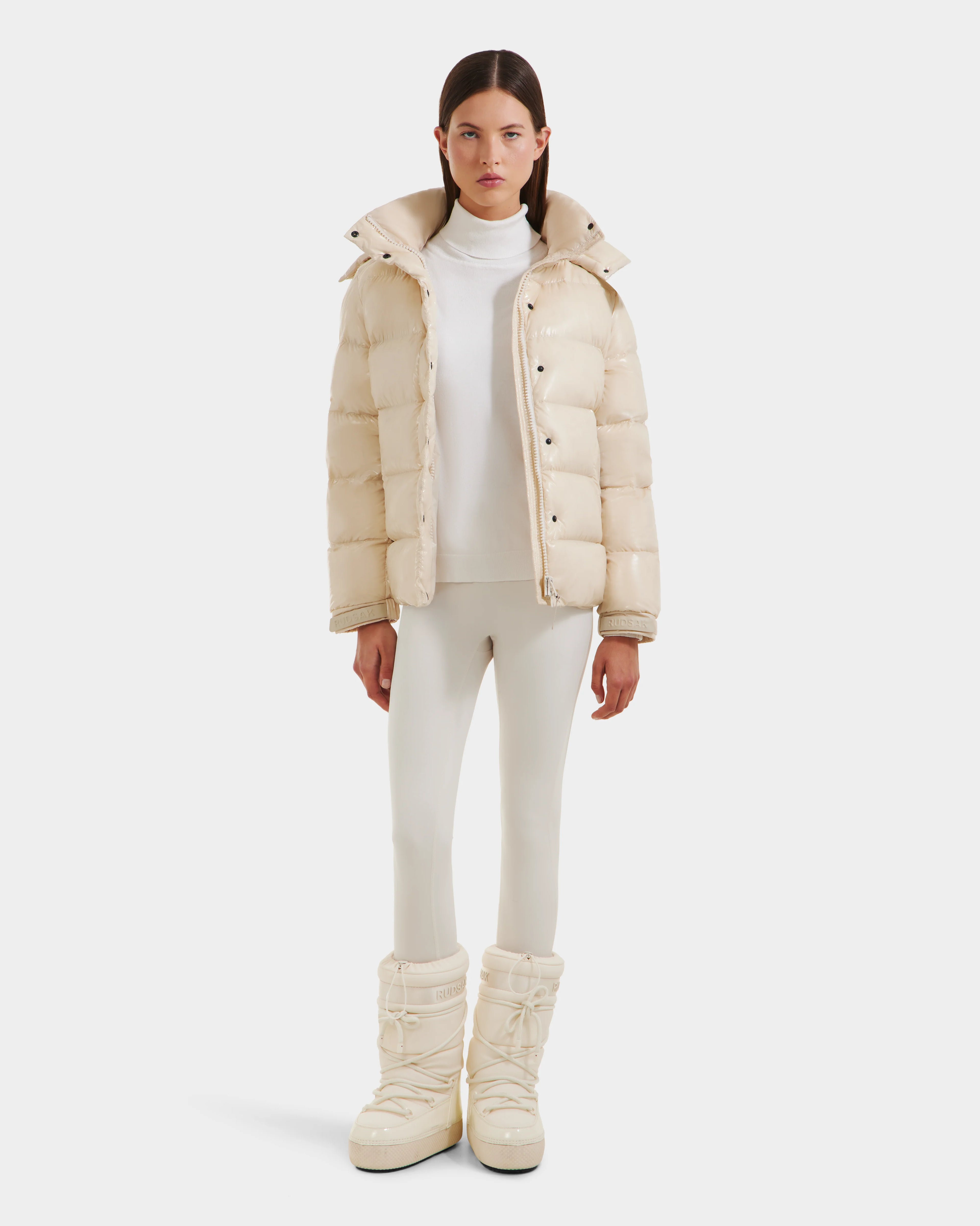 A woman with straight brown hair and a neutral expression stands facing forward against a white background, wearing the Rudsak BREE S - L125583 Vanilla women's down puffer with removable hood, cream snow boots, white turtleneck, and fitted white pants.