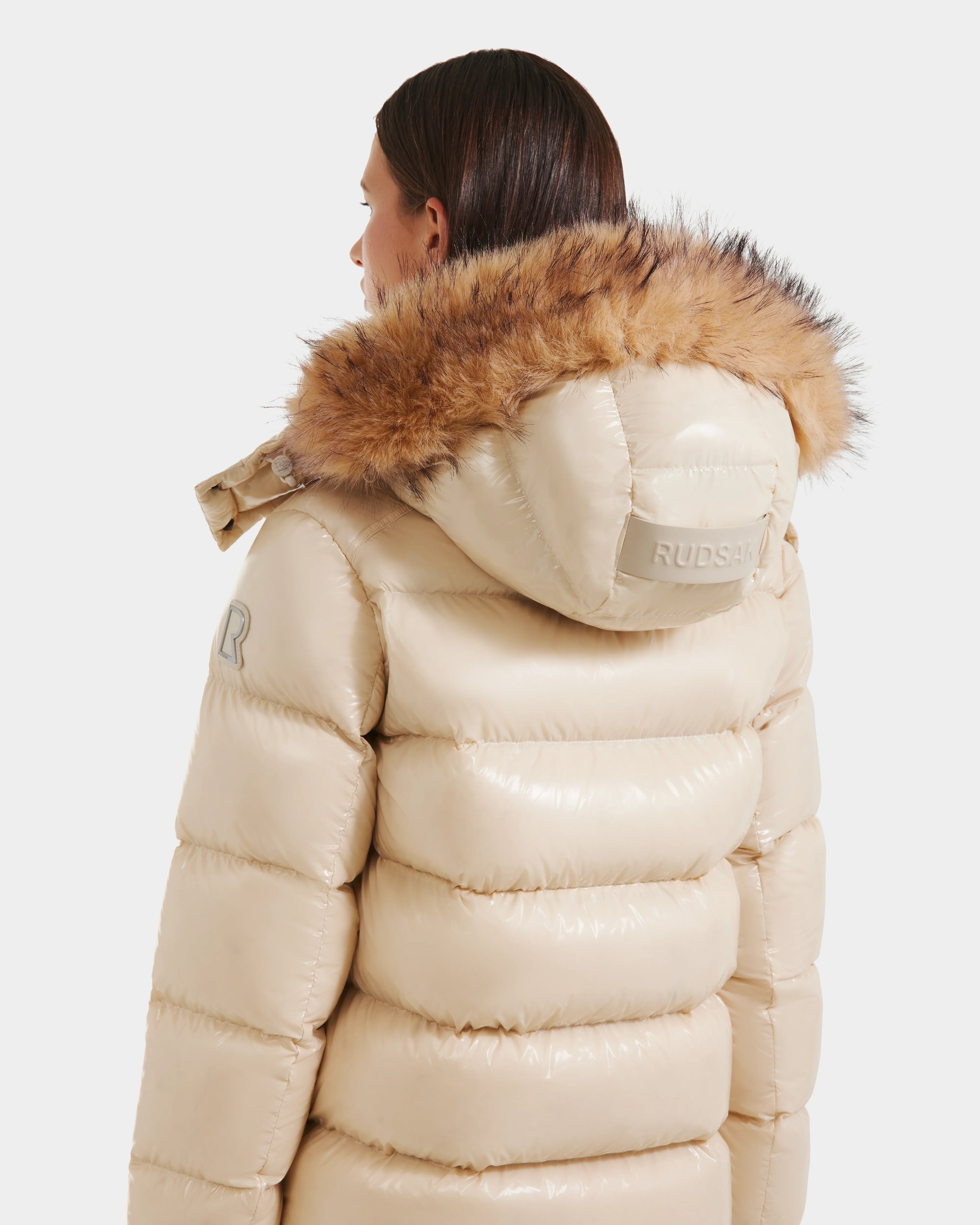 A woman with straight brown hair is seen from the back, wearing the Rudsak BREE S - L125583 down puffer jacket in Vanilla with a shiny ultra-gloss finish, a fur-trimmed removable hood, and Rudsak branding against a plain white background.