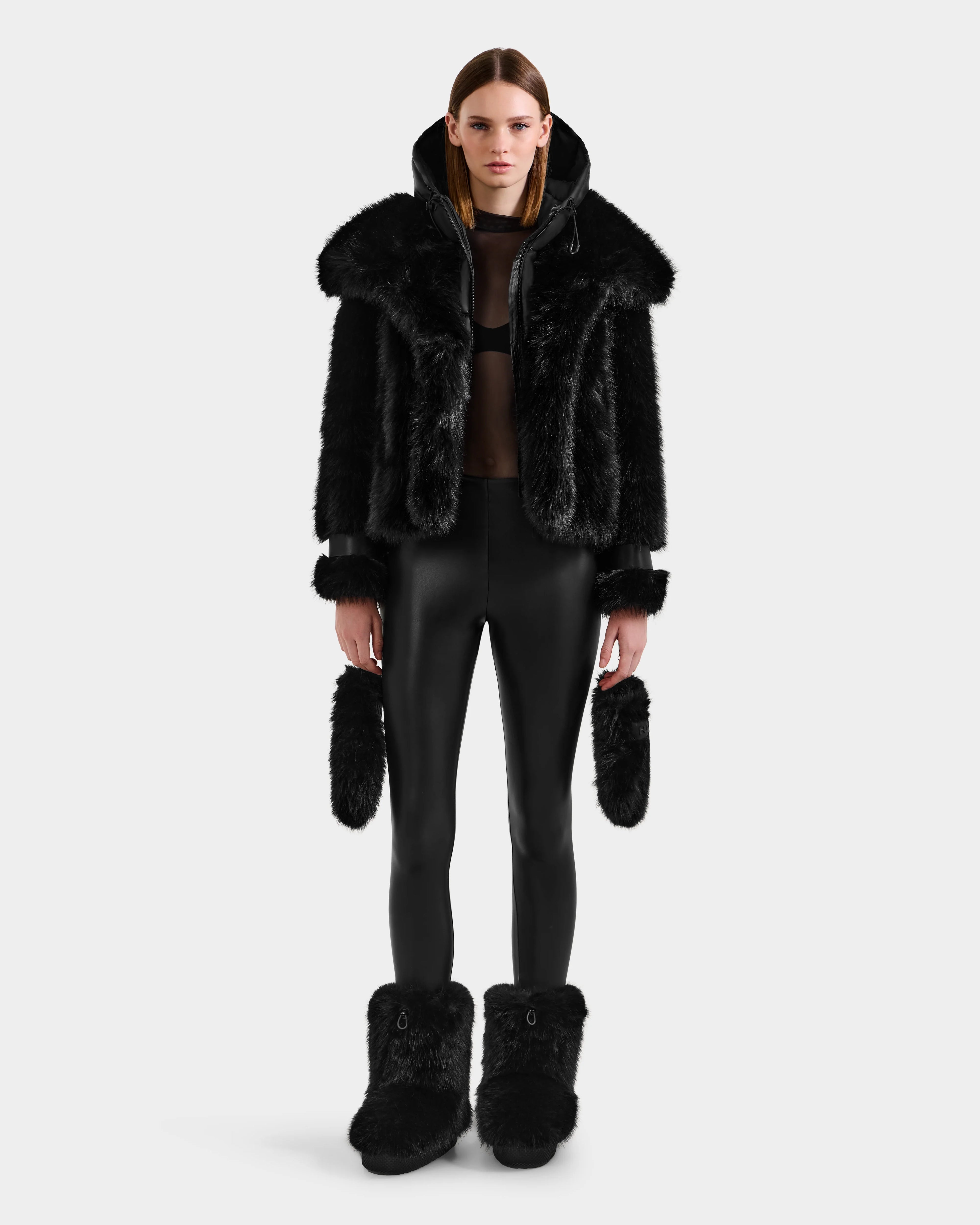 A person stands facing forward, wearing Rudsak's CHLOE - 8125619 Black women's faux fur jacket, with matching furry boots and mittens, black leggings, and a black top—an elevated winter look set against a plain white background.