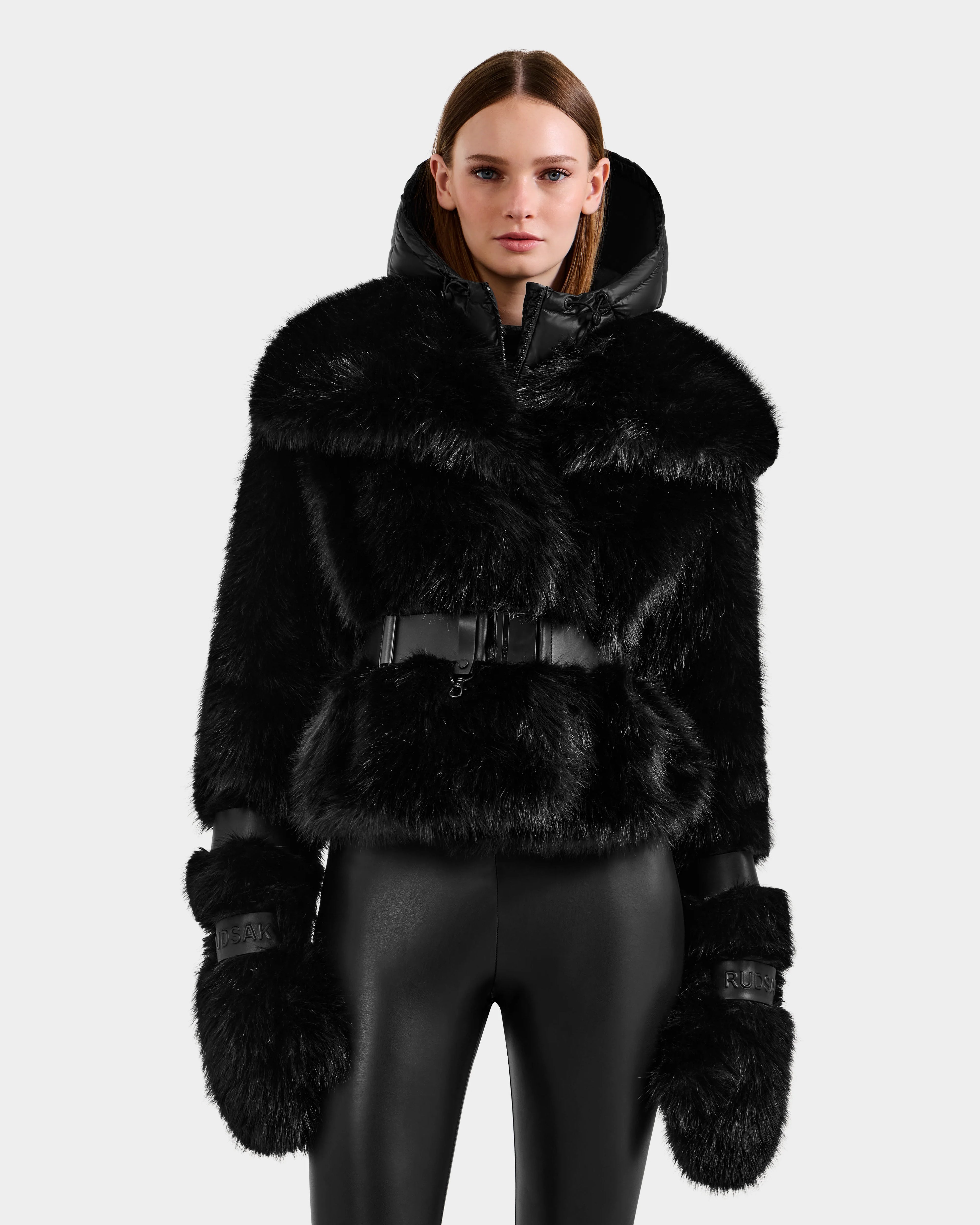A woman stands facing forward against a plain light background, wearing the CHLOE - 8125619 Black faux fur jacket and matching mittens by Rudsak, paired with shiny black pants for an effortlessly chic winter look.