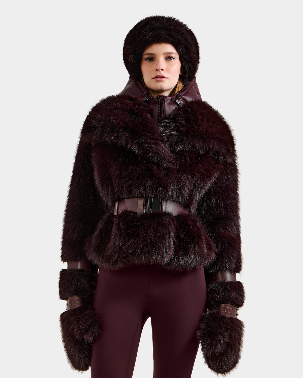 A person wearing the Rudsak CHLOE - 8125619 Bordeaux women's faux fur jacket with matching mittens and burgundy pants stands against a light background—a chic addition to any winter wardrobe.