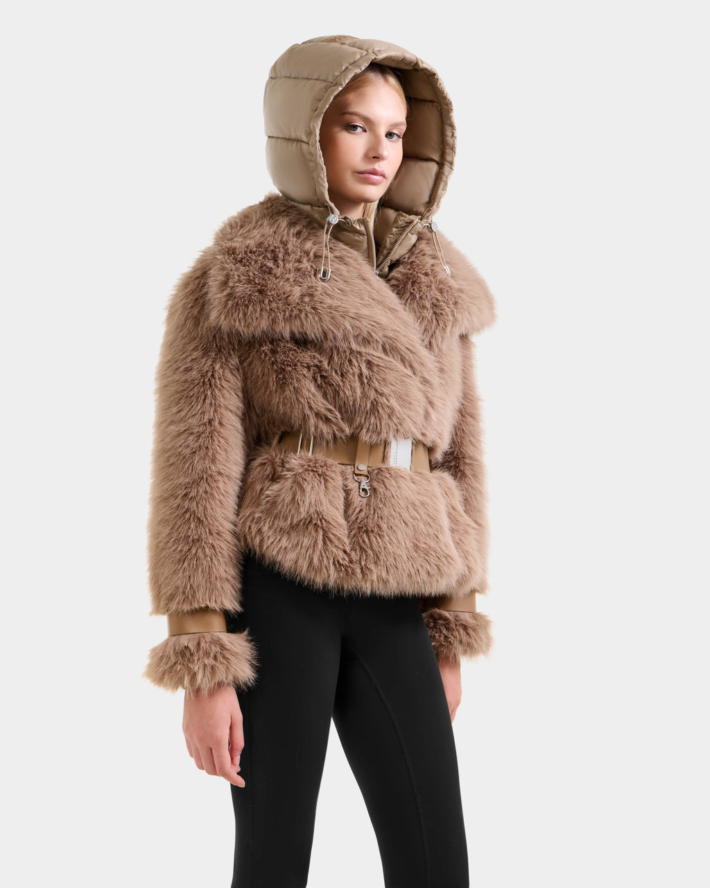 A person models the CHLOE - 8125619 Camel women's faux fur jacket by Rudsak, featuring a hood and beige belt, paired with black pants against a light gray background—a cozy choice for winter.