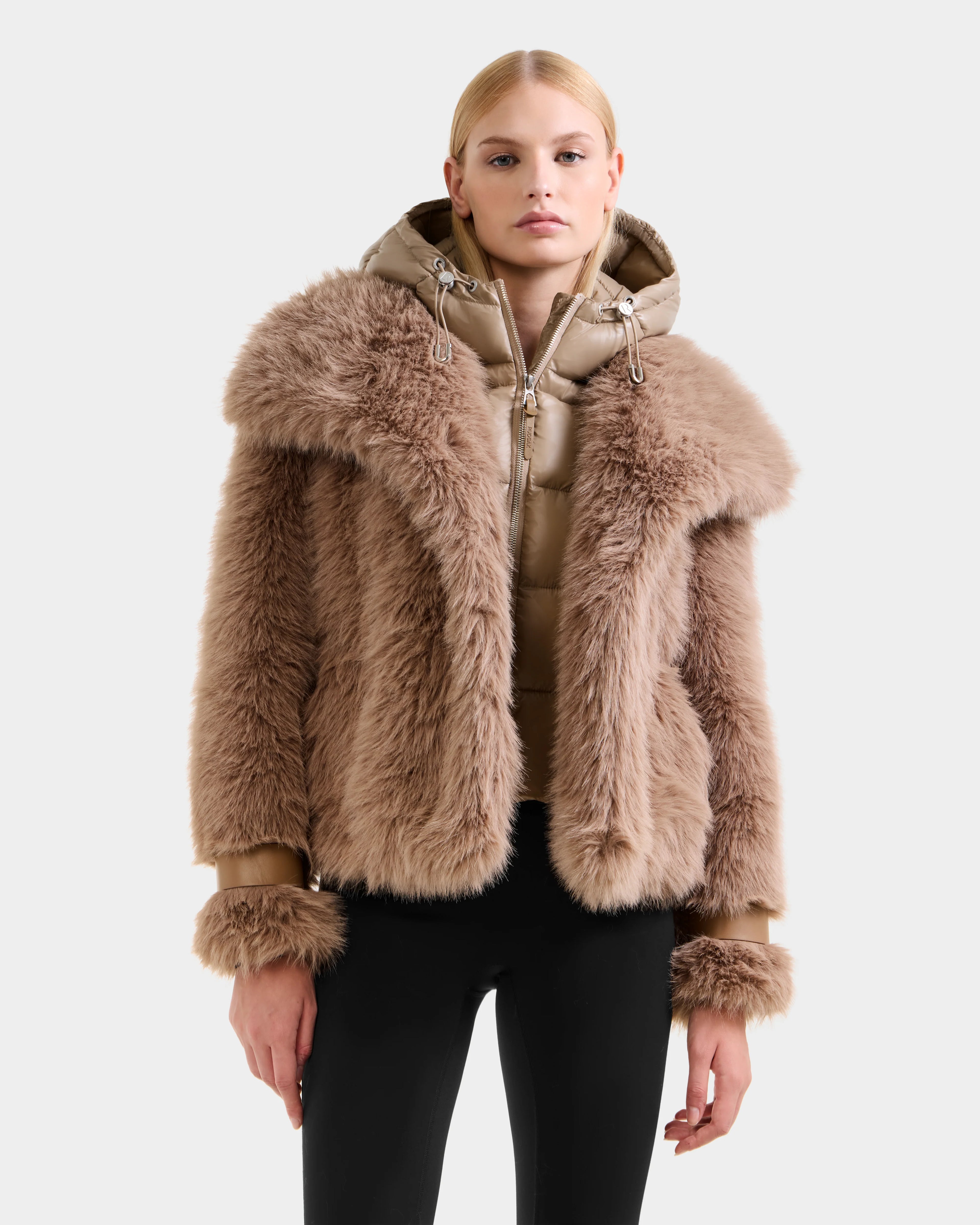A woman wears the Rudsak CHLOE - 8125619 Camel faux fur jacket with large collar and cuffs, layered over a hooded tan puffer, paired with black fitted pants—an essential winter look against a plain light gray background.