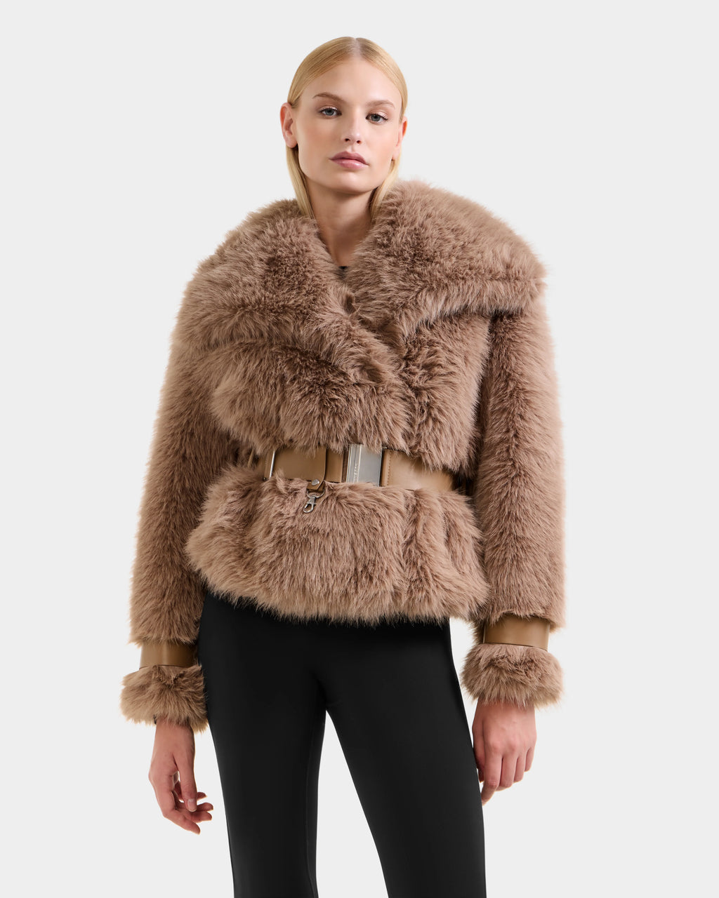 A woman models the Rudsak CHLOE - 8125619 Camel faux fur jacket with a wide collar and belt, paired with black leggings against a light gray background—an essential piece for any winter wardrobe.