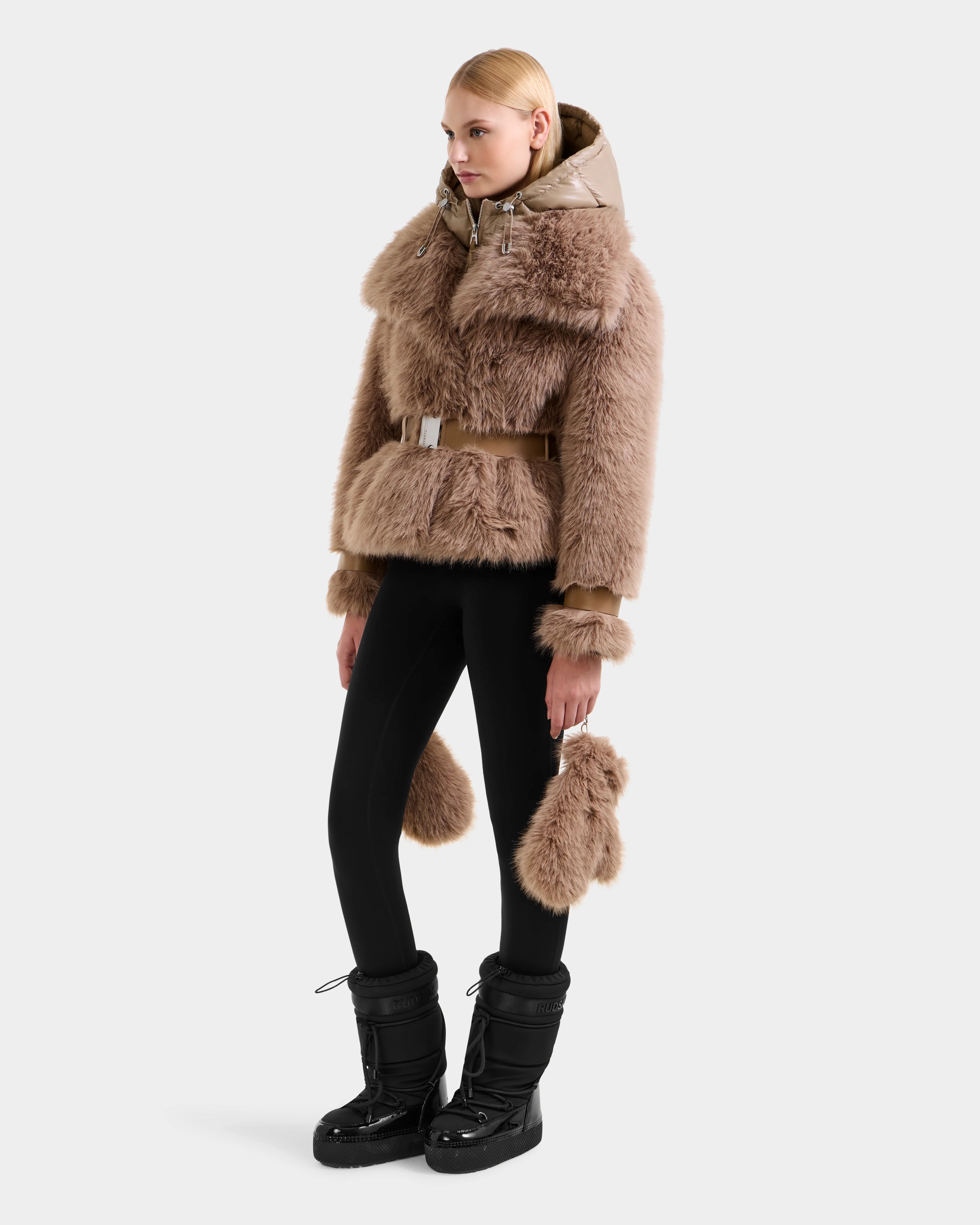 A woman with straight blonde hair stands sideways in a Rudsak CHLOE - 8125619 Camel faux fur belted jacket, black leggings, and winter boots, holding a matching tan accessory—an elegant winter look against a plain white background.