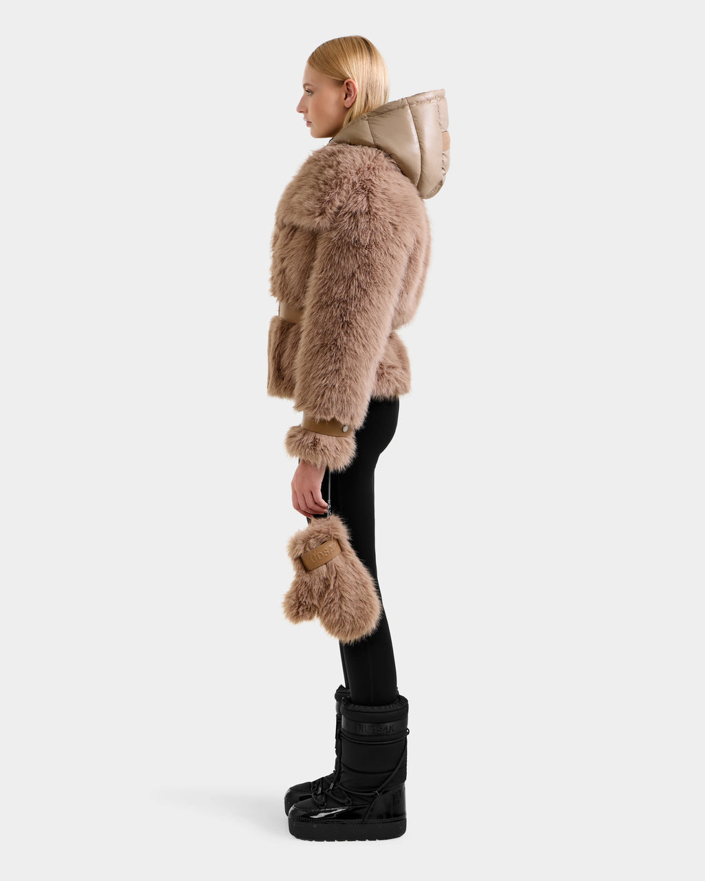 A person in profile models the Rudsak CHLOE - 8125619 Camel women's faux fur jacket with shiny beige hood, paired with black leggings, black winter boots, and a matching chic faux fur accessory against a plain white background.