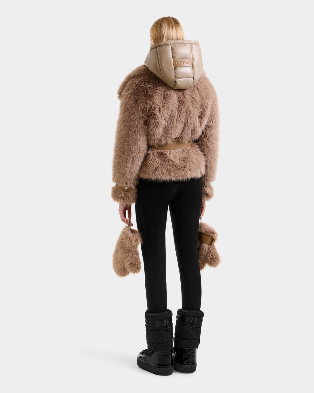 A person wearing the Rudsak CHLOE - 8125619 Camel faux fur jacket with hood, black leggings, and snow boots holds a matching accessory while facing away, highlighting a chic winter look against a plain white background.