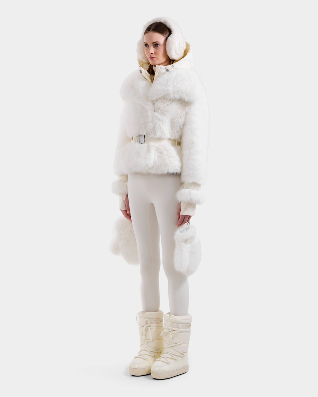 A woman models the Rudsak CHLOE - 8125619 Iceland white faux fur hooded jacket with matching earmuffs, fitted white pants, and cream snow boots for an effortlessly chic winter look against a plain white background.