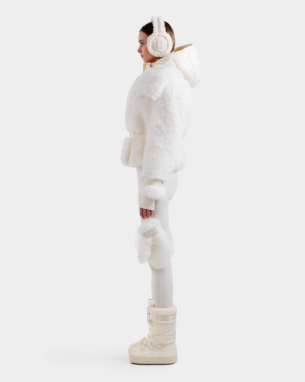 A woman stands in profile wearing the Rudsak CHLOE - 8125619 Iceland faux fur jacket with a fluffy white hood, paired with white fitted pants, furry mittens, and cream winter boots—showcasing chic style against a light background.