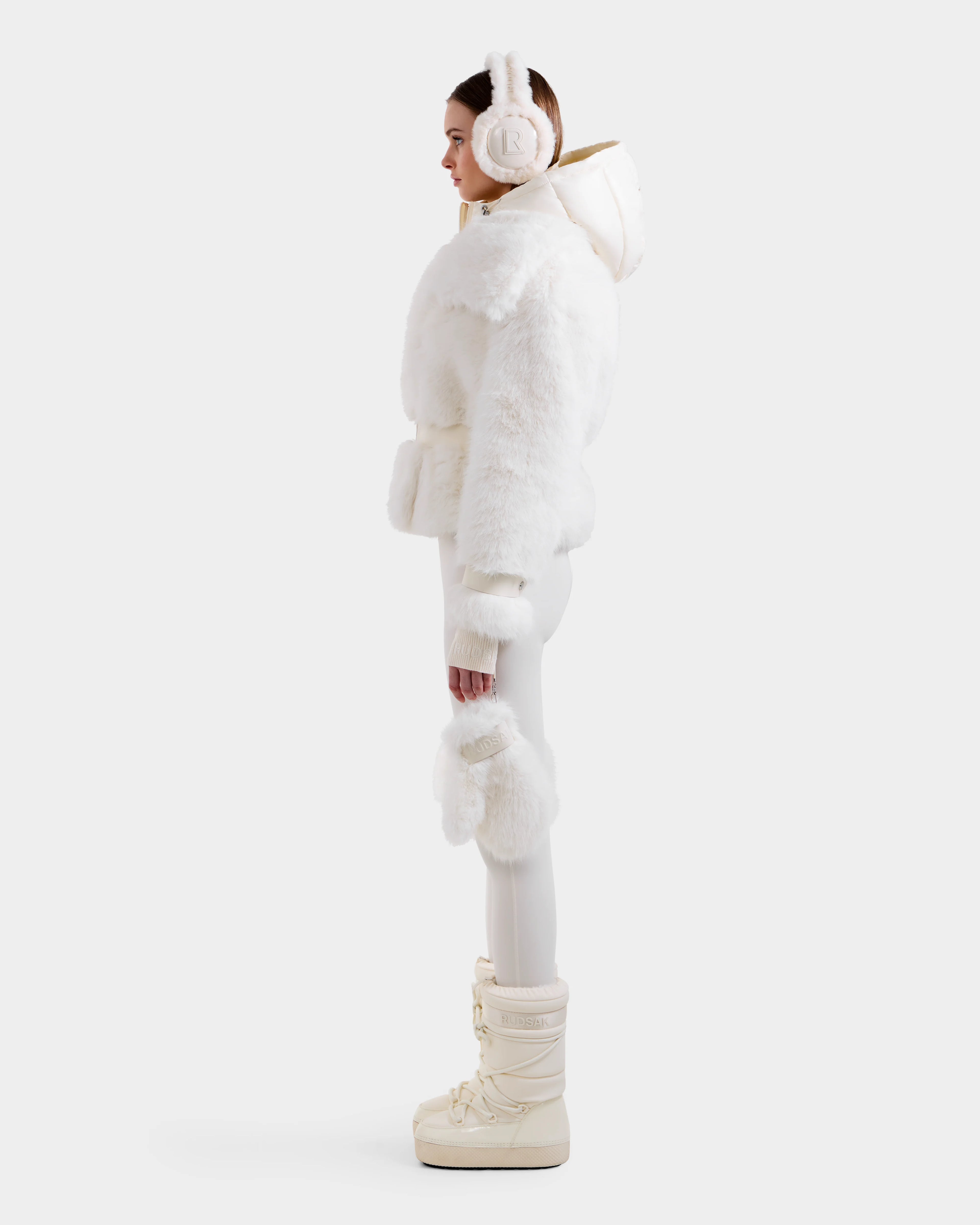 A woman stands in profile wearing the Rudsak CHLOE - 8125619 Iceland faux fur jacket with a fluffy white hood, paired with white fitted pants, furry mittens, and cream winter boots—showcasing chic style against a light background.