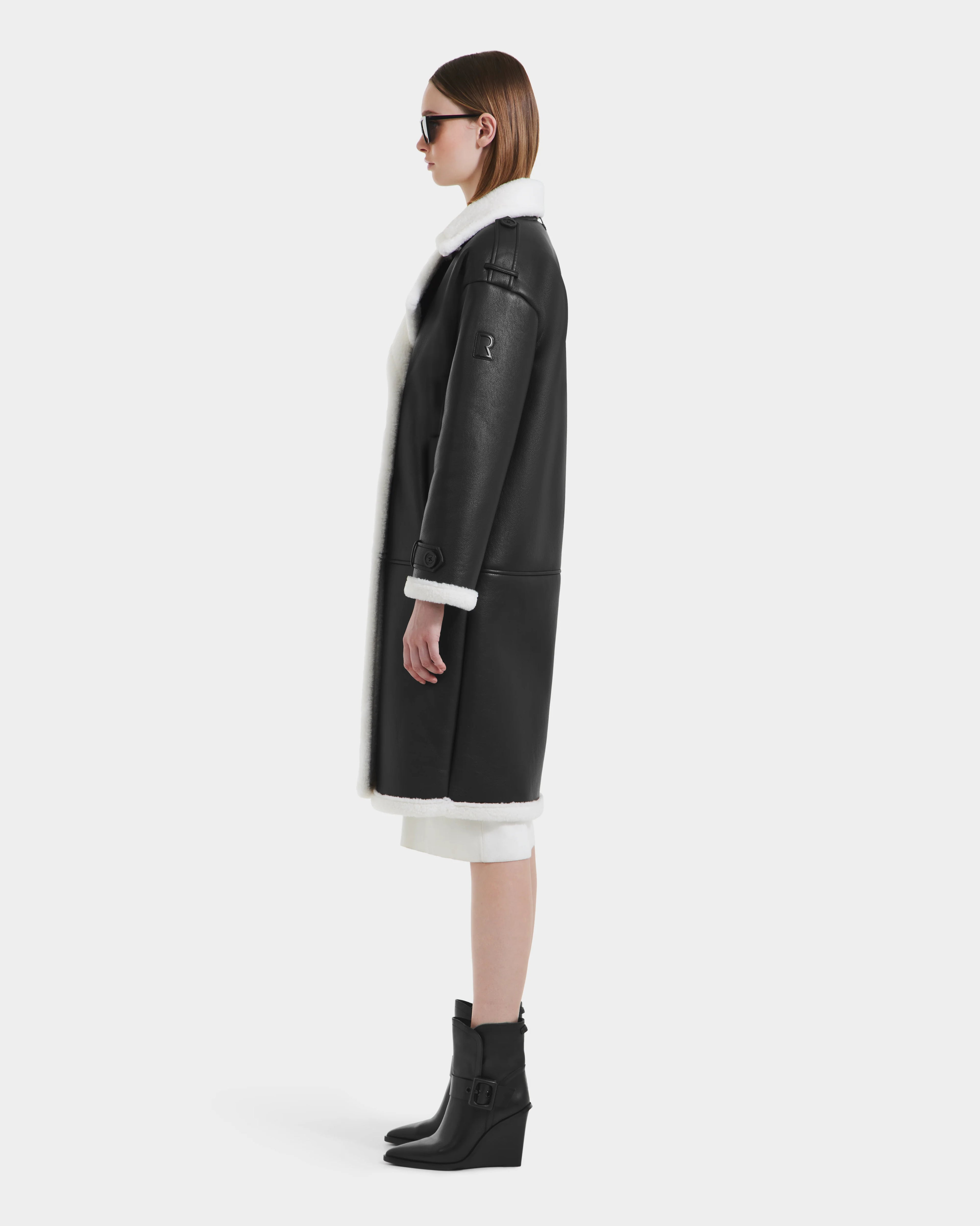 A woman in sunglasses models the Rudsak DARCY - L125539 Black women's winter coat with white lining and black ankle boots, standing in profile against a plain white background.