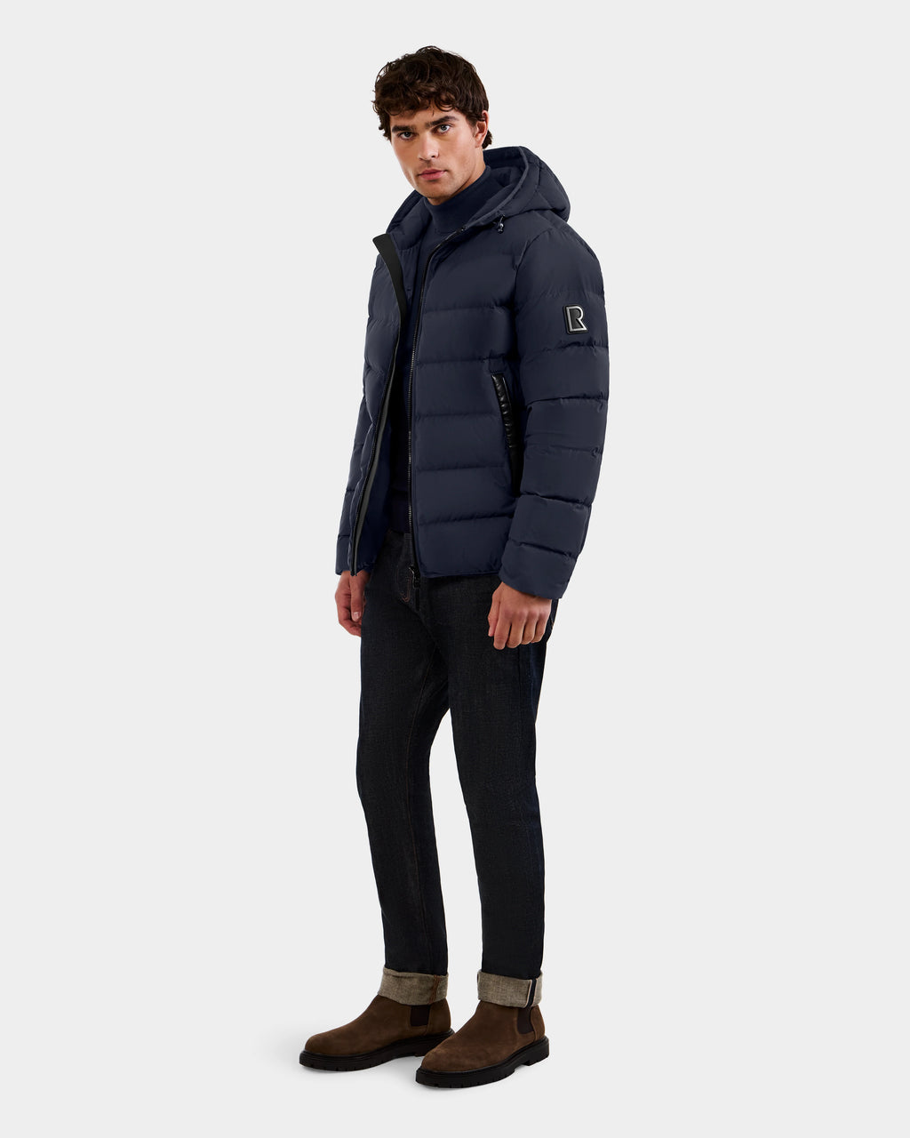 A man stands facing forward in the Rudsak DEMIAN M - M123507 Navy men’s down puffer jacket, made from RDS-certified premium down and recycled fabric. He pairs it with dark cuffed jeans and brown suede boots against a plain light gray background.