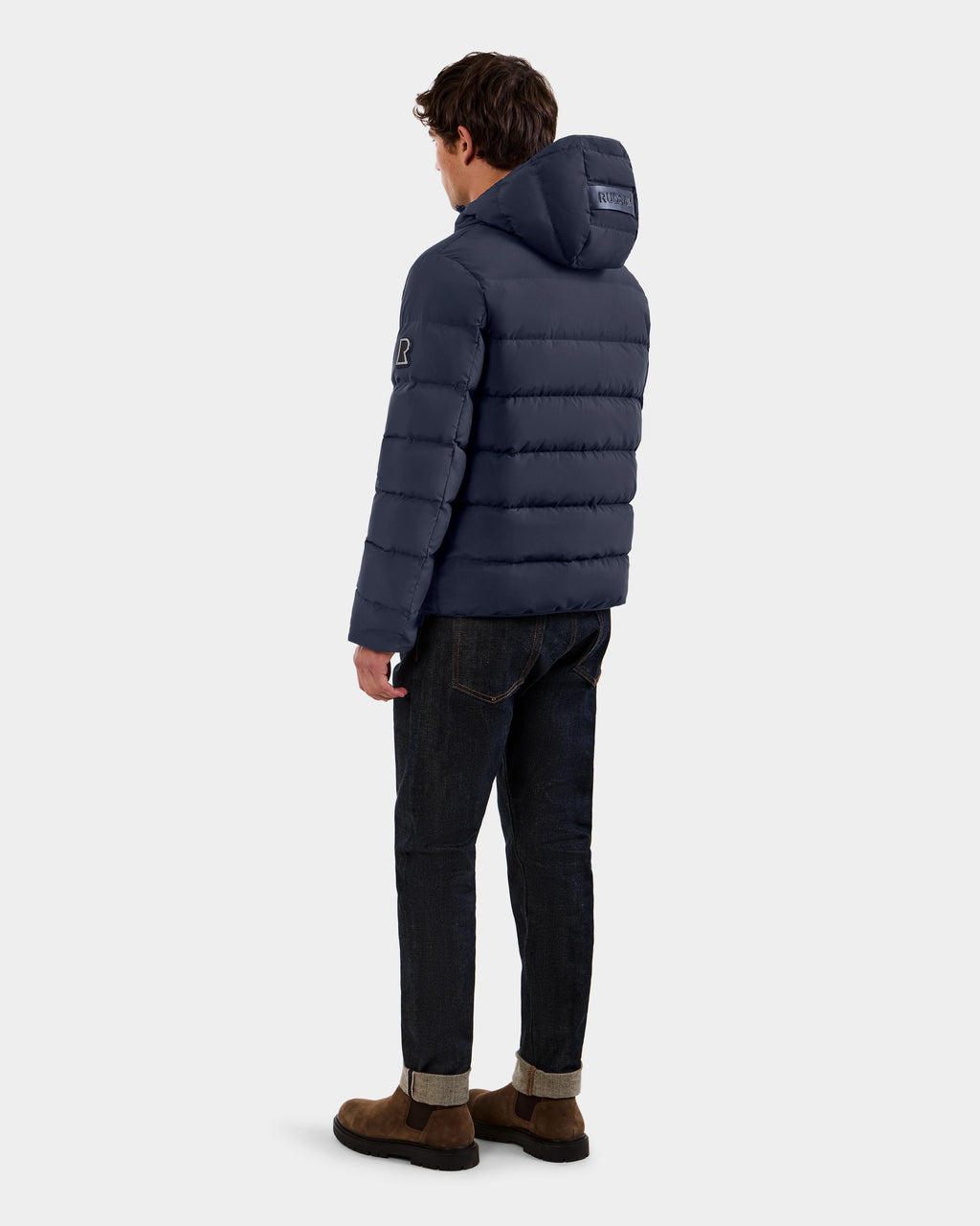 A man stands facing away, wearing the Rudsak DEMIAN M - M123507 navy hooded down puffer made from sustainable recycled fabric, paired with dark jeans and brown suede shoes against a plain light background.