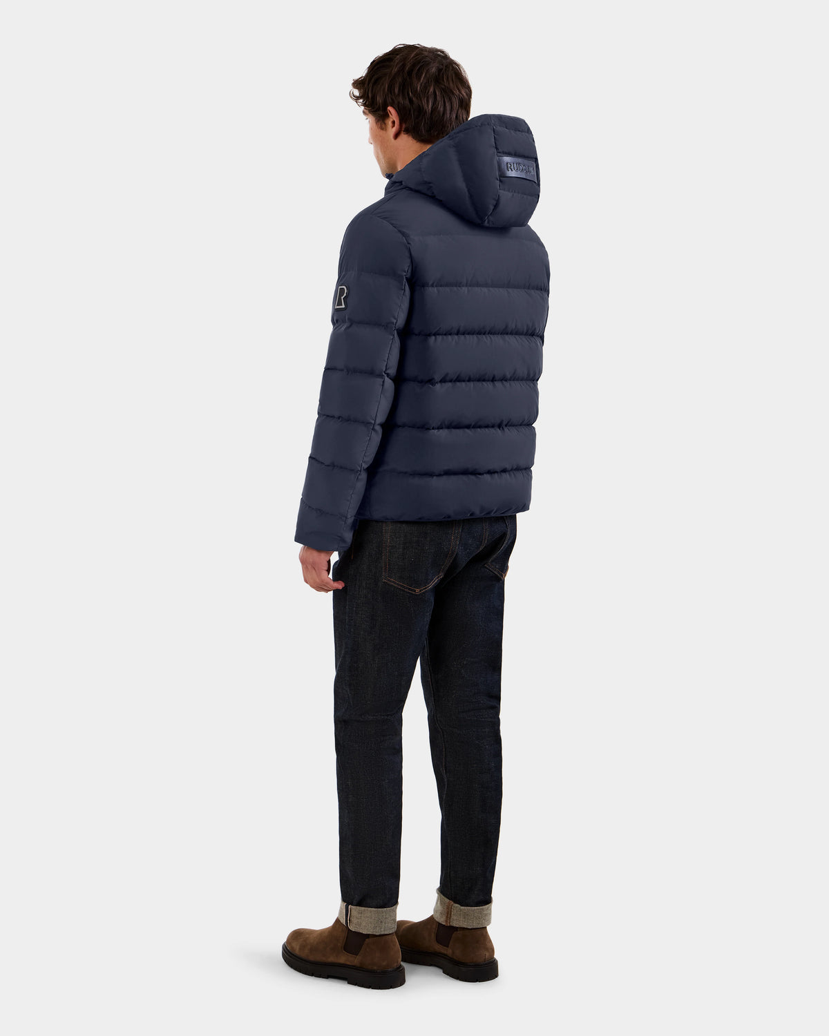 A man stands facing away, wearing the Rudsak DEMIAN M - M123507 navy hooded down puffer made from sustainable recycled fabric, paired with dark jeans and brown suede shoes against a plain light background.