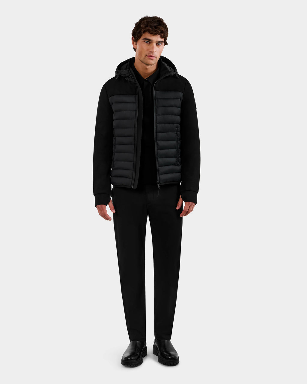 A man stands facing forward wearing the Rudsak DURIN M - M125530 Black men’s hooded jacket, paired with black pants and black shoes, against a plain light gray background.