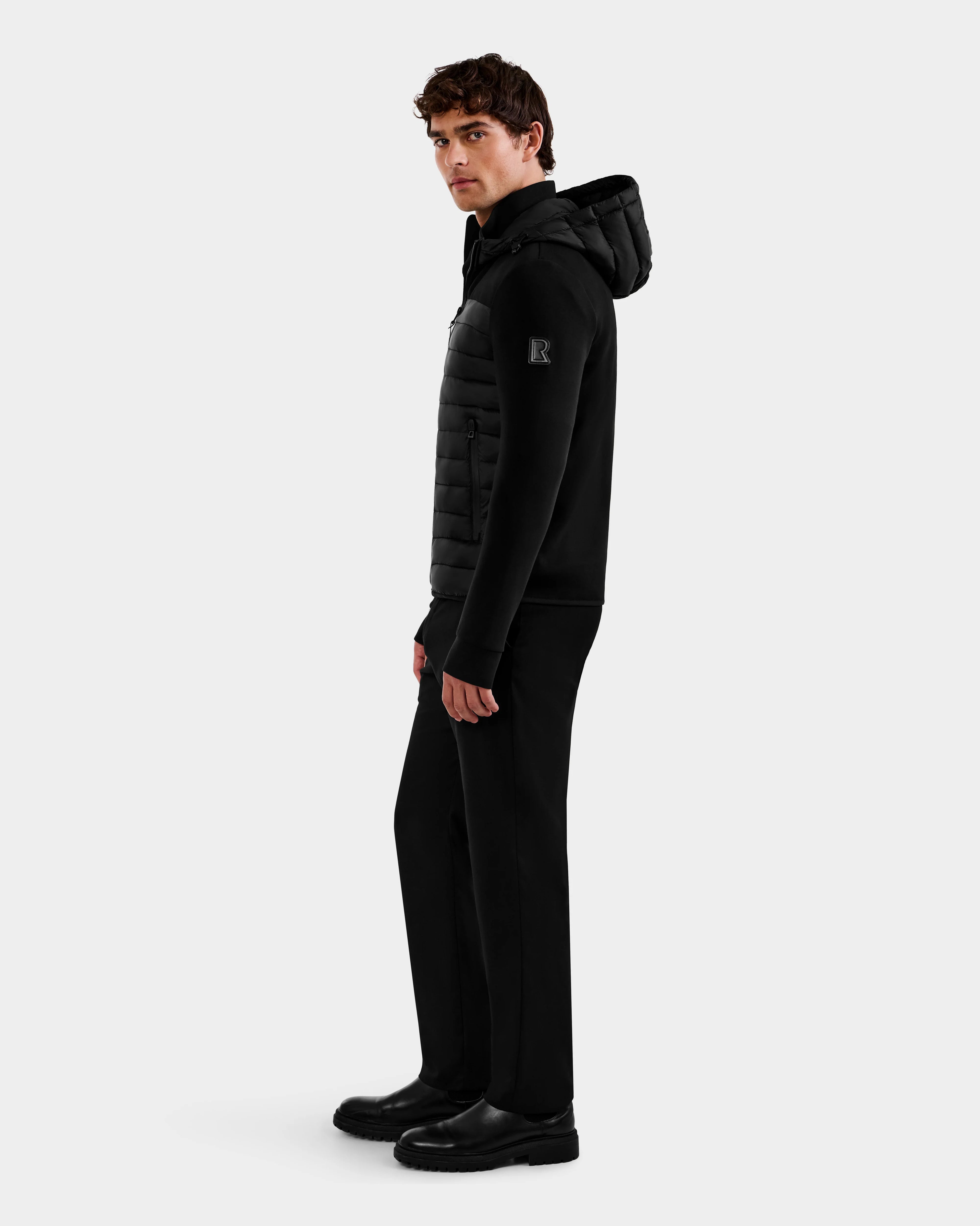 A young man stands on a light gray background wearing the DURIN M - M125530 Black men’s hooded jacket with waterproof zippers by Rudsak, paired with straight pants and boots. He is facing sideways but looking toward the camera.