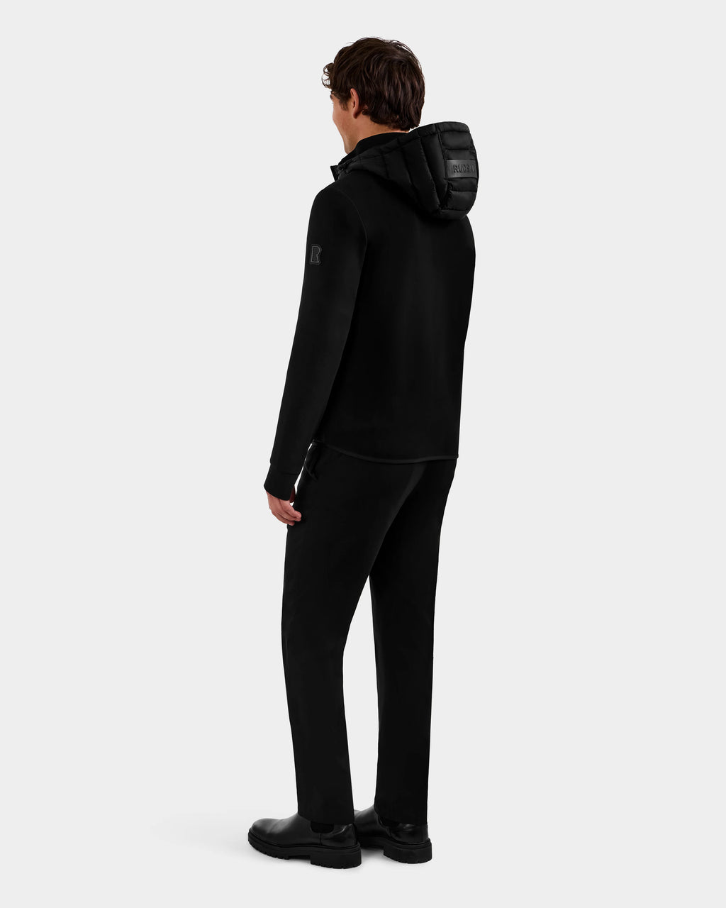 A man stands facing away wearing the Rudsak DURIN M - M125530 Black men’s hooded jacket with waterproof zippers, paired with black pants and shoes, set against a light gray background.