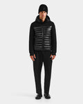 A man stands facing forward against a plain light gray background, wearing the Rudsak DURIN S - M125529 Black men's hooded jacket with horizontal panels and lightweight down insulation, along with black pants, beanie, and shoes.