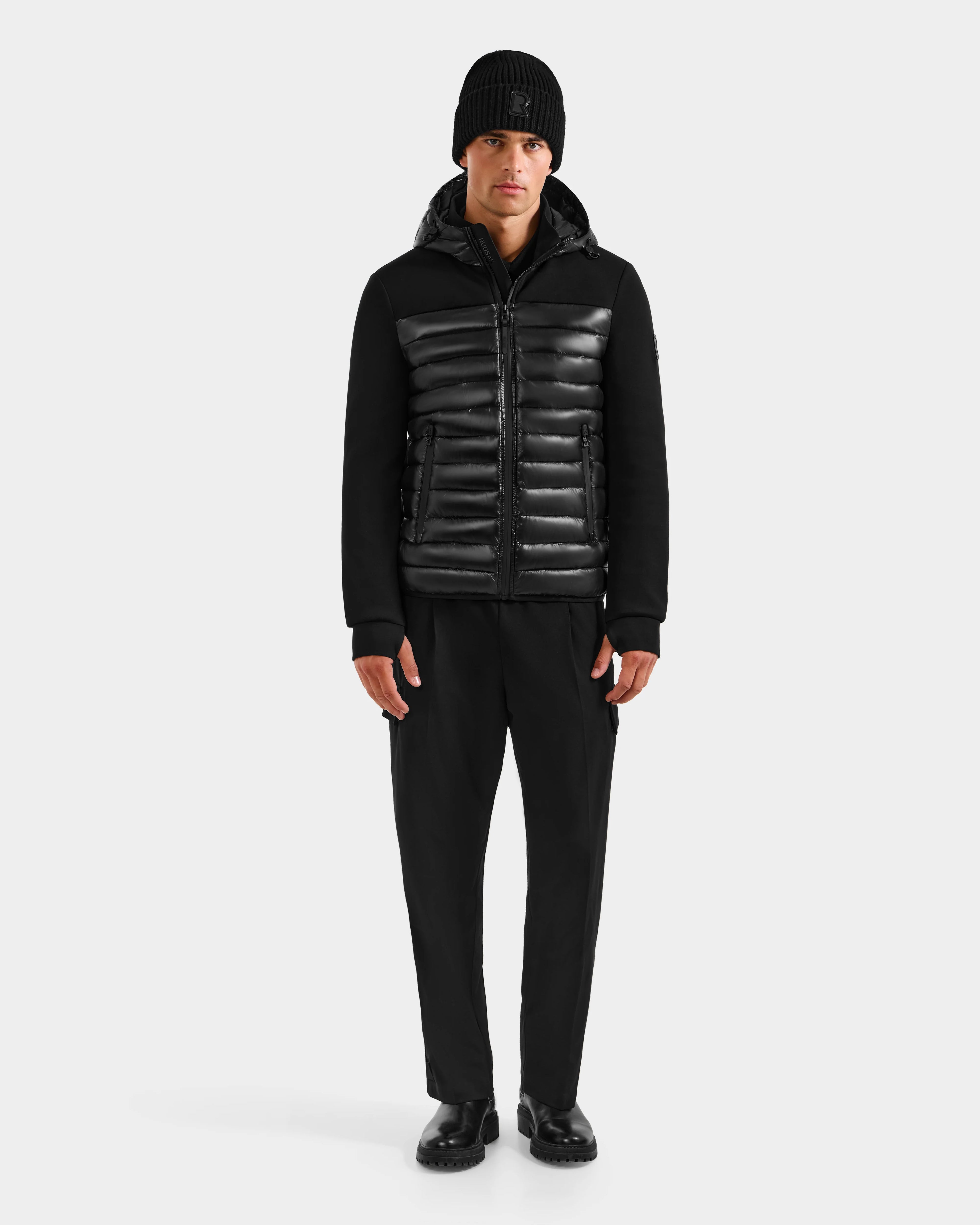 A man stands facing forward against a plain light gray background, wearing the Rudsak DURIN S - M125529 Black men's hooded jacket with horizontal panels and lightweight down insulation, along with black pants, beanie, and shoes.