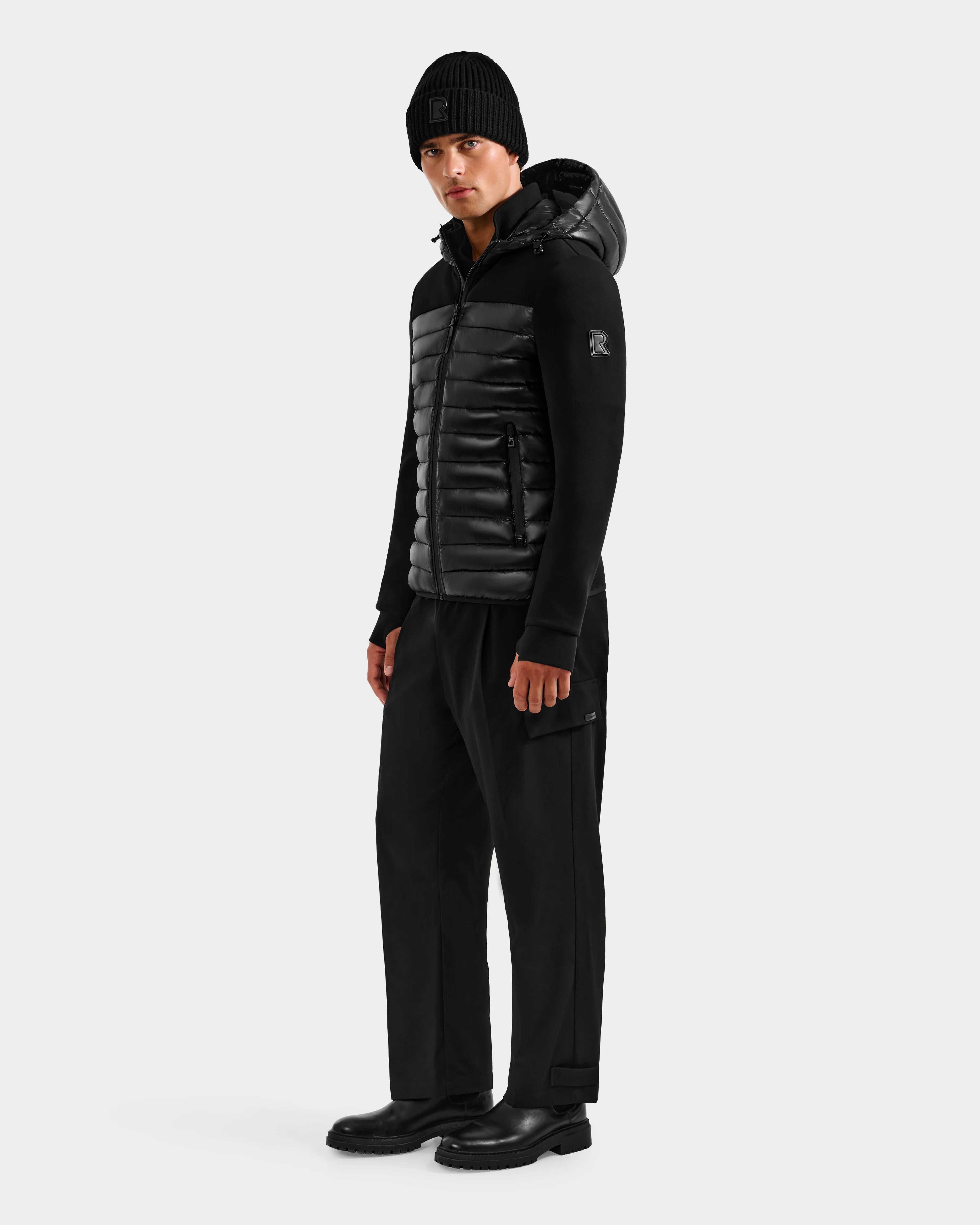 A man in the Rudsak DURIN S - M125529 Black hooded jacket stands facing slightly to the side, dressed in black pants, black boots, and a black knit beanie against a plain light gray background.