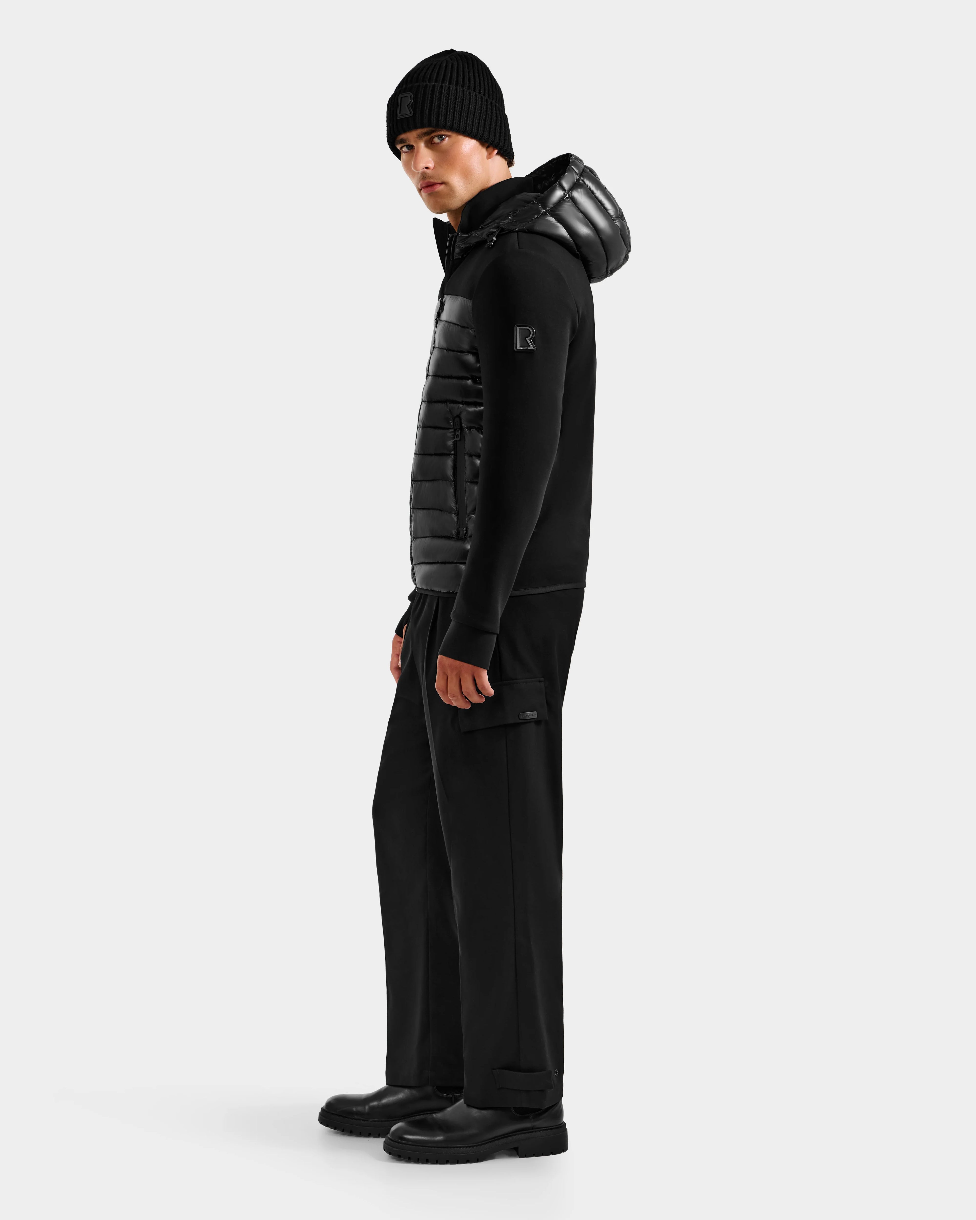 A man faces slightly left, wearing a Rudsak DURIN S - M125529 Black men’s hooded jacket with lightweight down insulation, black cargo pants, and black boots against a plain light background.