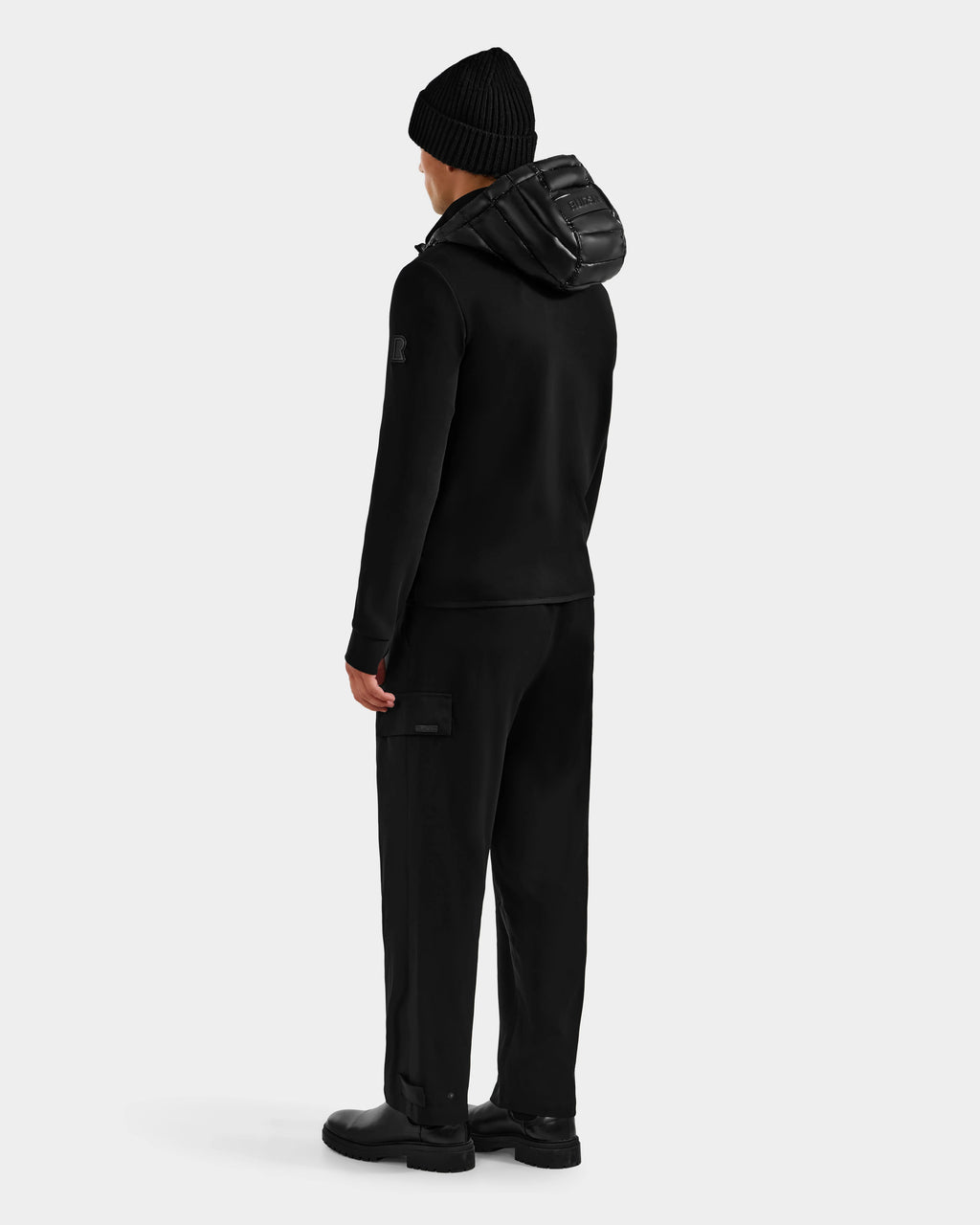 A person wearing the Rudsak DURIN S - M125529 Black men’s hooded jacket stands facing away, showcasing its back, along with black pants and boots, against a plain white background.