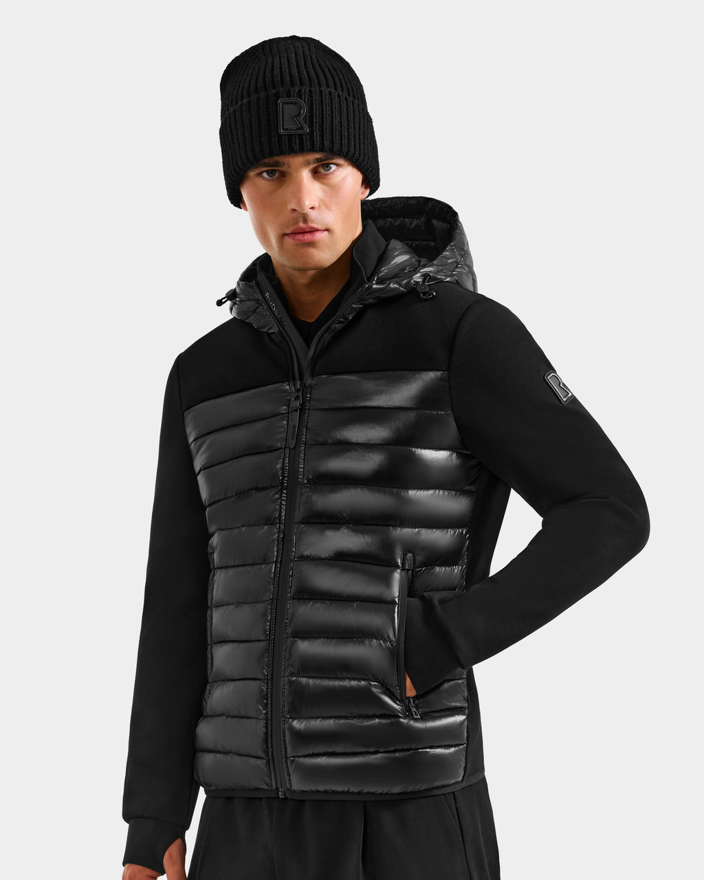A person wearing the Rudsak DURIN S - M125529 Black men’s hooded jacket with horizontal quilting, waterproof zippers, and black pants poses against a plain light gray background.