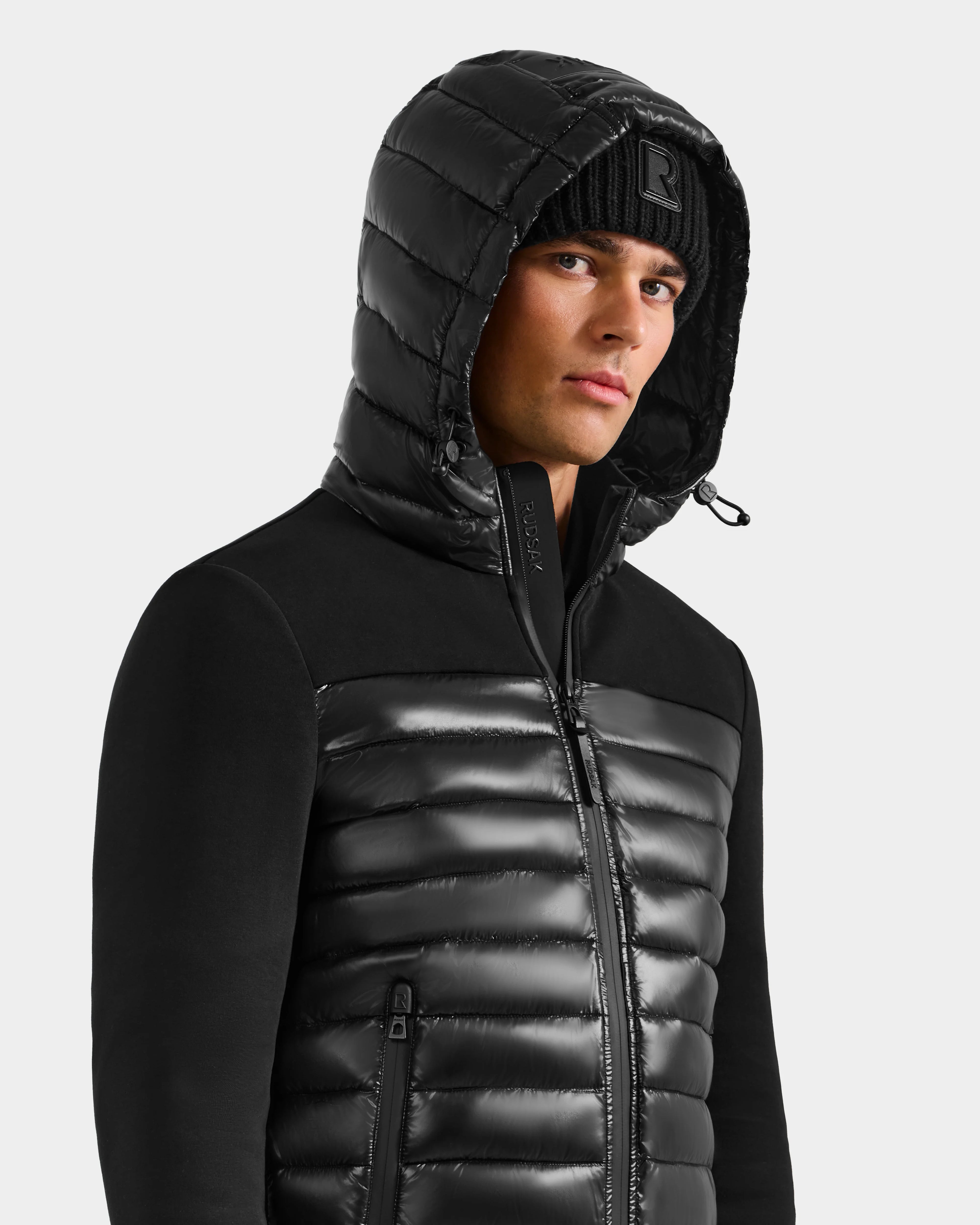 A man in a shiny black Rudsak DURIN S - M125529 men’s hooded jacket with lightweight down insulation and a black knit beanie, looking slightly to the side against a plain light background.