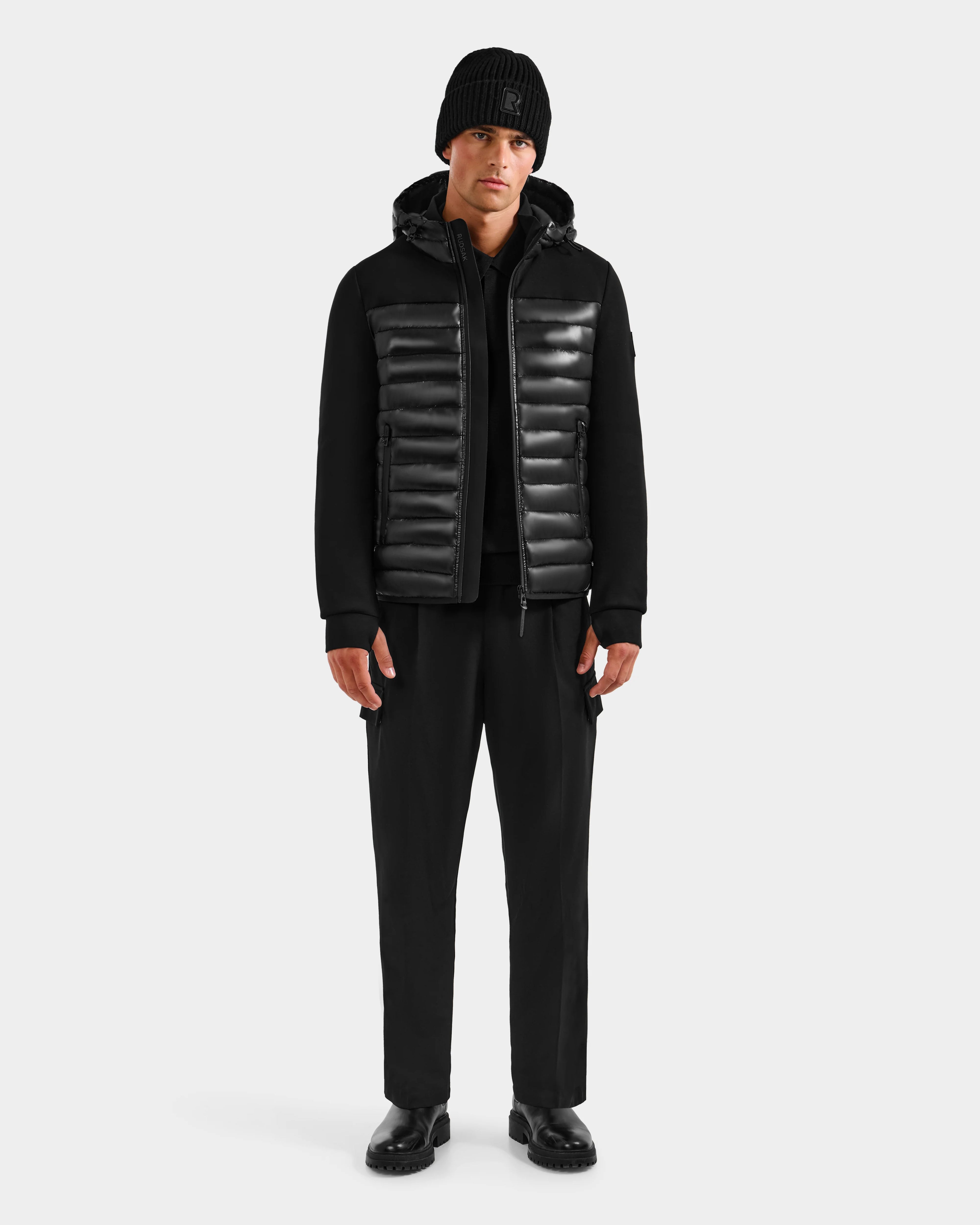 A man wearing Rudsak’s DURIN S - M125529 Black men’s hooded jacket with black pants. The jacket offers lightweight down insulation for warmth and features waterproof zippers for protection.
