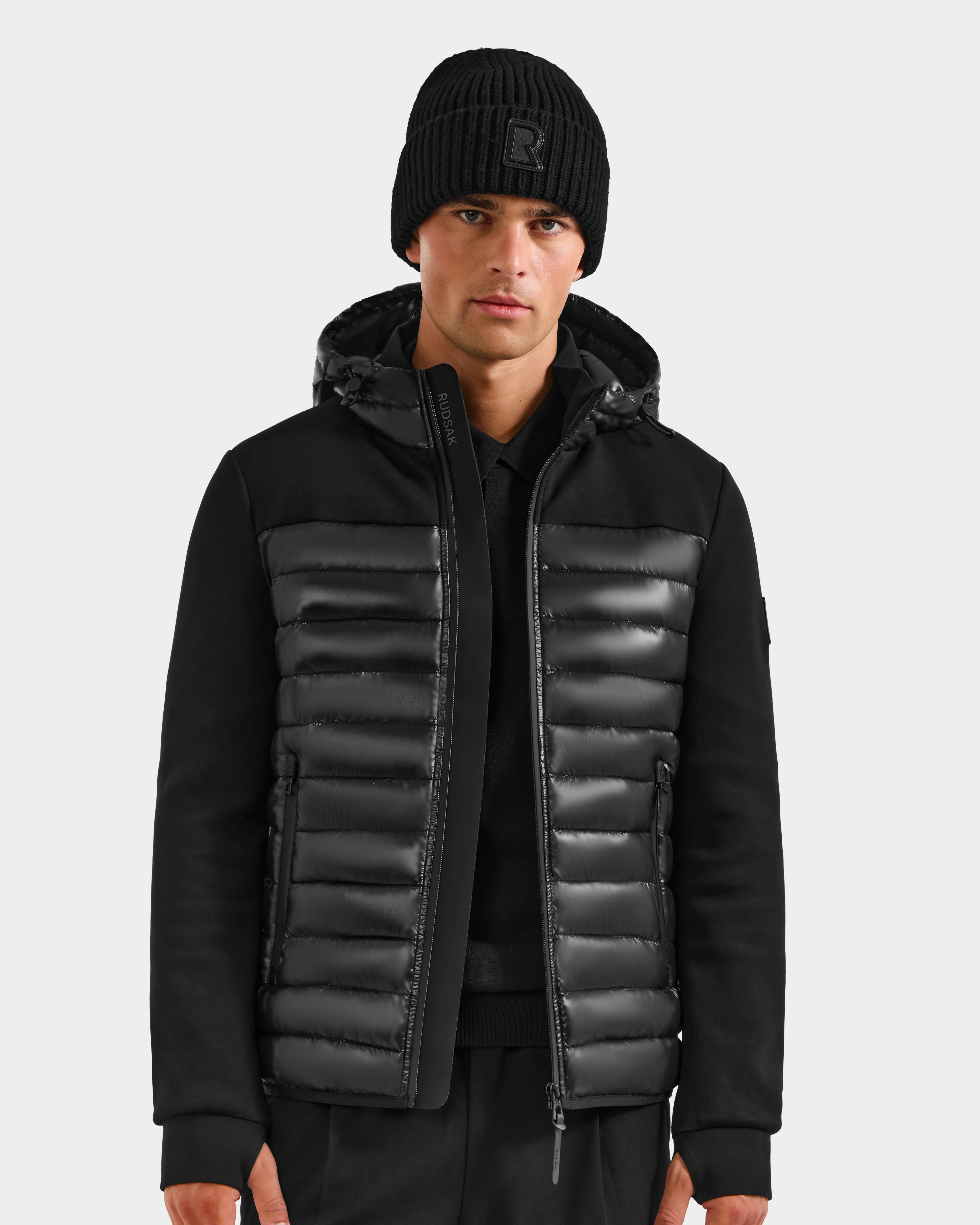 A man wears the Rudsak DURIN S - M125529 Black padded men’s hooded jacket with a shiny quilted front, lightweight down insulation, and waterproof zippers, paired with a black knit beanie and pants against a plain light background.