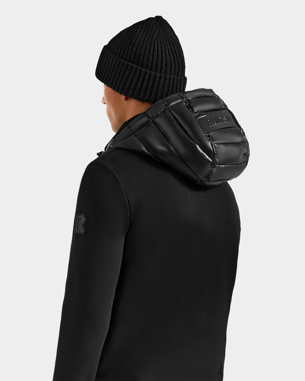 A person is shown from behind, wearing the Rudsak DURIN S - M125529 Black men’s hooded jacket with lightweight down insulation and a shiny, padded hood, set against a plain white background.
