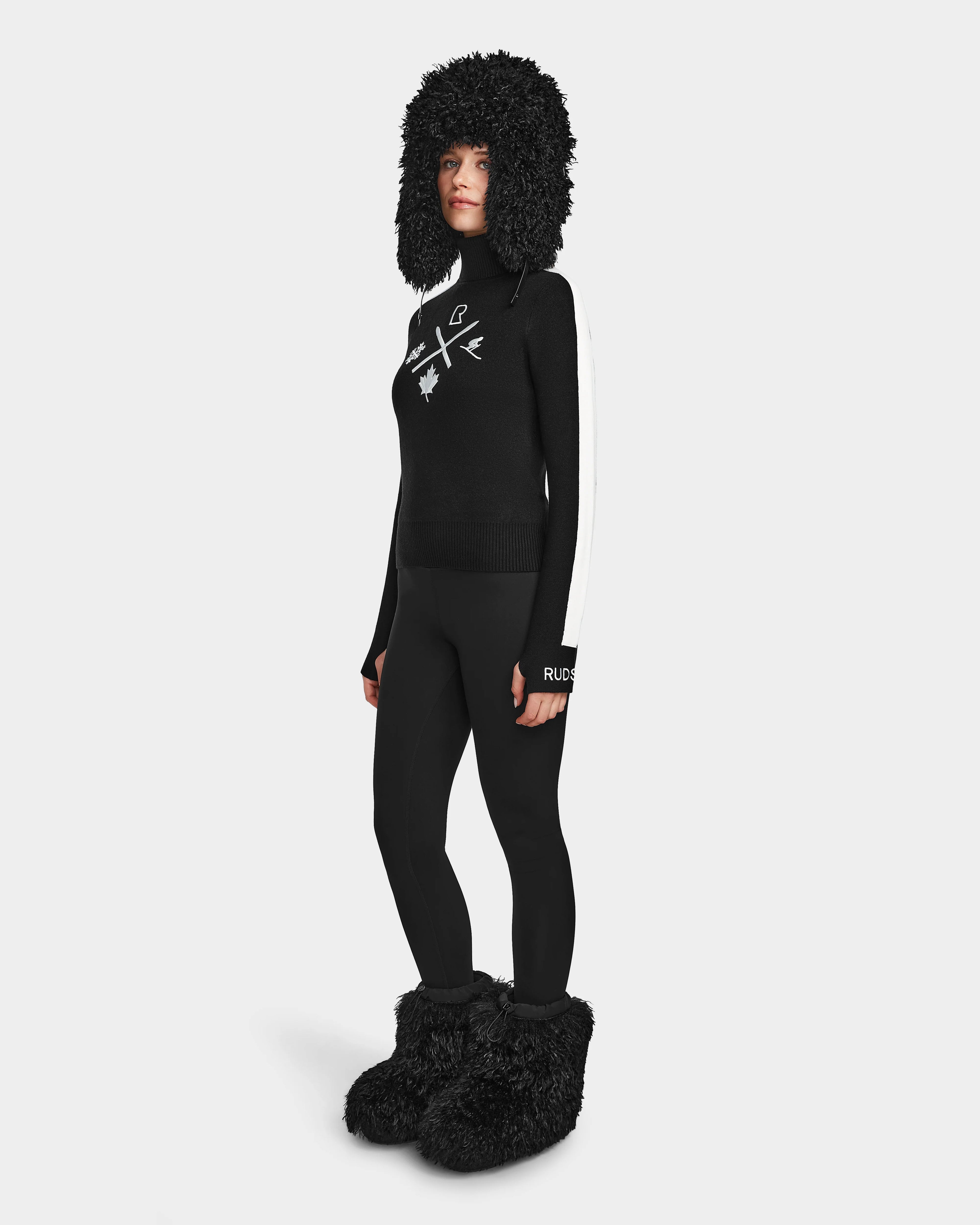 A person wears the Rudsak ELIZABETH - 8524623 Black, styled with a black furry hat, womens ski turtleneck with white abstract designs, black leggings, and oversized furry boots—an ideal après-ski winter look against a plain white background.