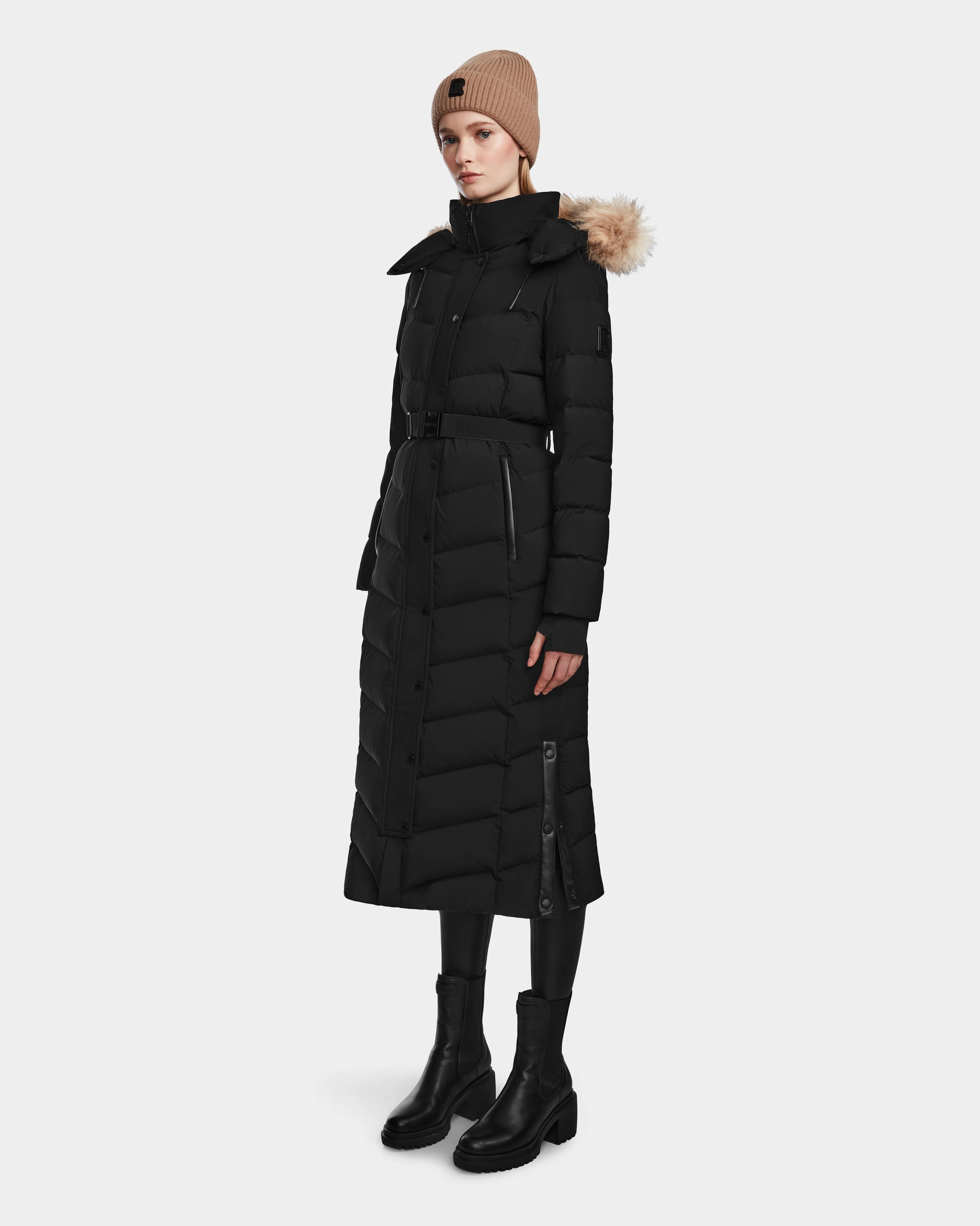 A woman wearing the Rudsak ELSIE M - L123517 Black/Natural maxi down puffer with a fur-trimmed hood, black boots, black pants, and a beige knit beanie stands against a plain light background.