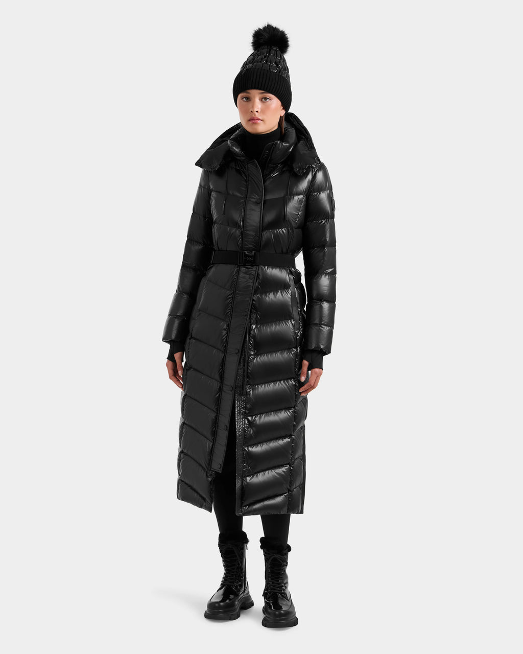 A person stands against a plain light background wearing the Rudsak ELSIE S - L124536 Black women’s maxi down puffer with water-resistant, RDS-certified down, paired with a black pom-pom knit hat, black pants, and lace-up boots.