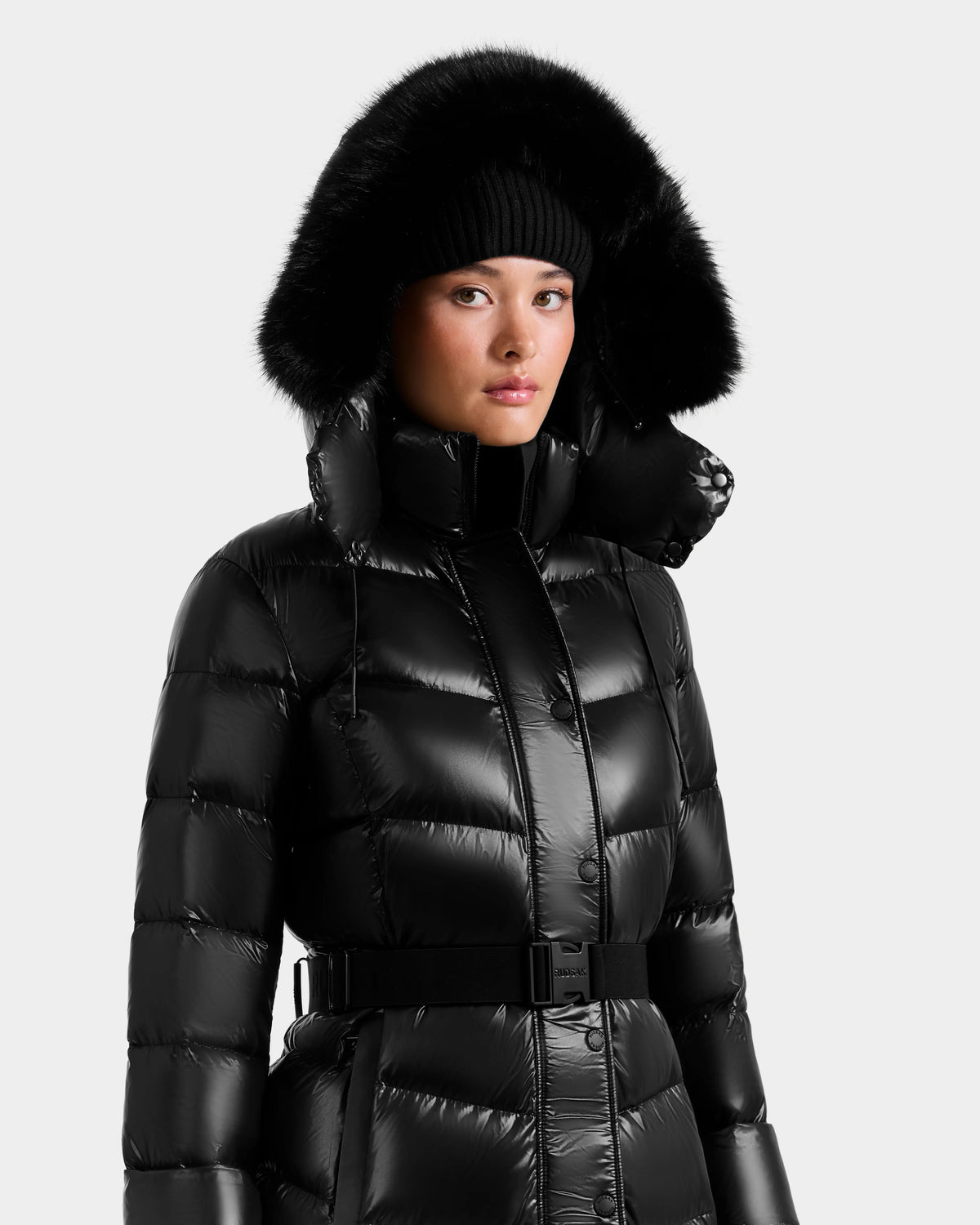 A person wears the Rudsak ELSIE S - L124536 Black women’s maxi puffer coat with a belted waist, high collar, and black fur-trimmed hood, paired with a black knit beanie against a plain, light background.