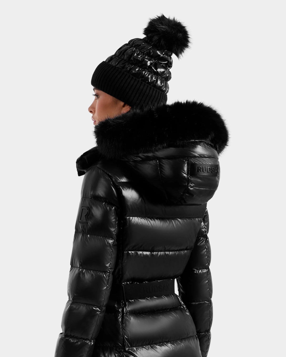 A person models the Rudsak ELSIE S - L124536 Black women’s maxi down puffer with a fur-trimmed hood and a black knit pom-pom beanie, viewed from the back and turned slightly to the side against a plain background.