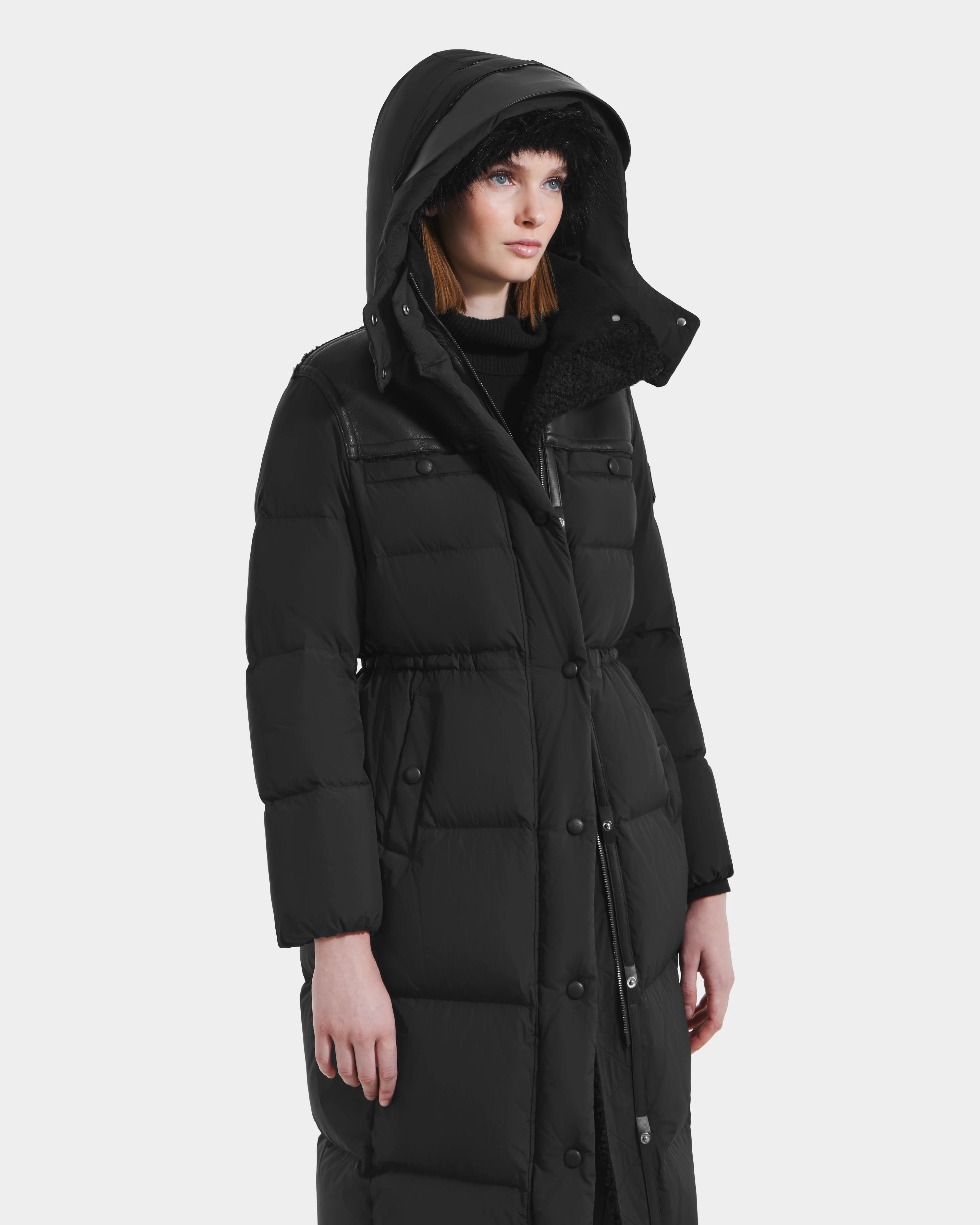 A woman stands slightly left against a white background, wearing the Rudsak EMBER M-L - L125543 Black women's long down puffer coat with a large removable hood and snap buttons.