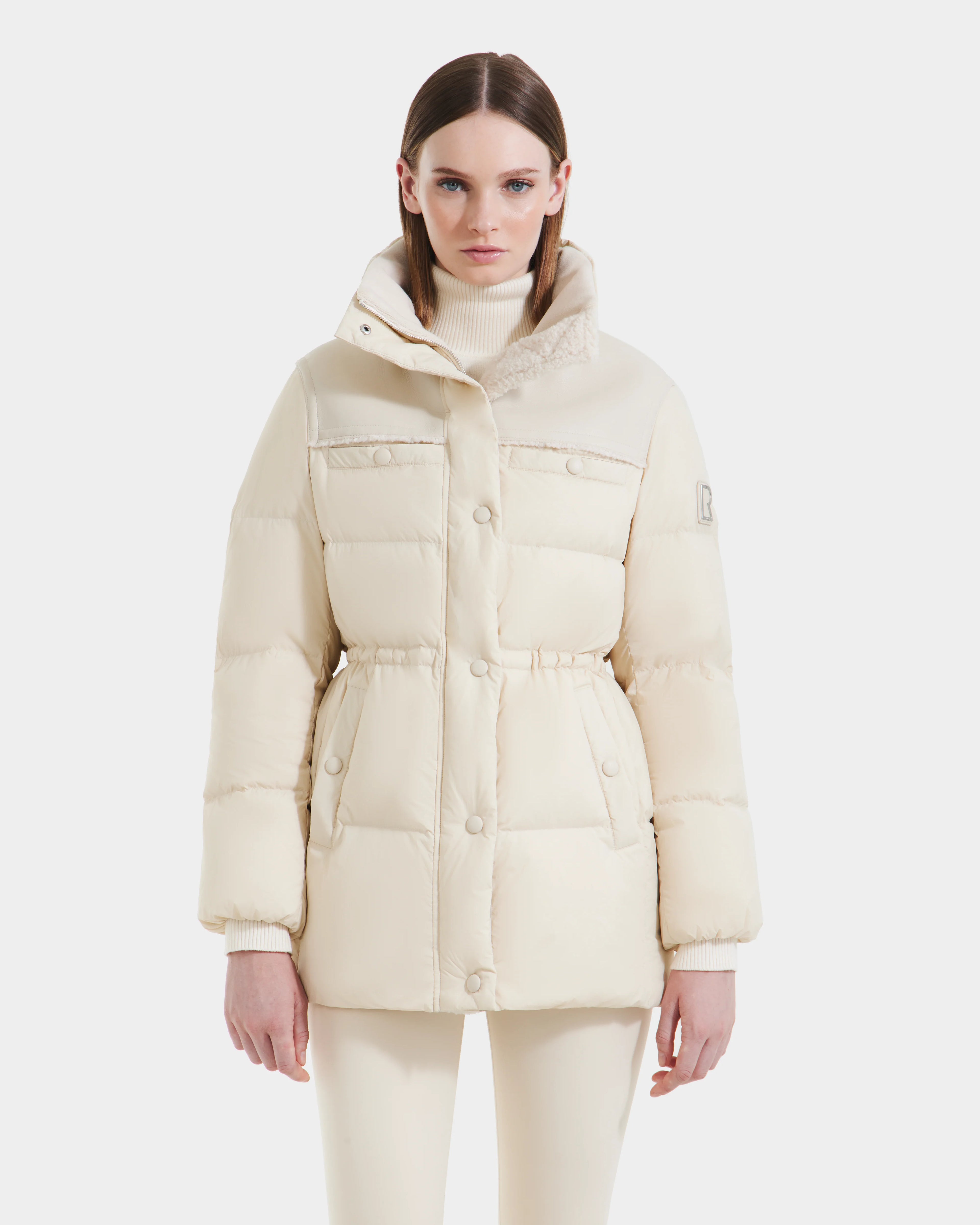 A woman in the Rudsak EMBER M-M - L125524 Vanilla light beige down puffer jacket with a high collar and front buttons, paired with matching beige pants and a turtleneck, stands against a plain white background.