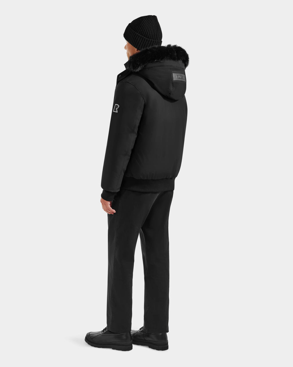 A person stands facing away, wearing the Rudsak JEROME - 6125514 Black men’s down bomber jacket with fur trim, a black beanie, black pants, and black shoes against a plain light gray background.