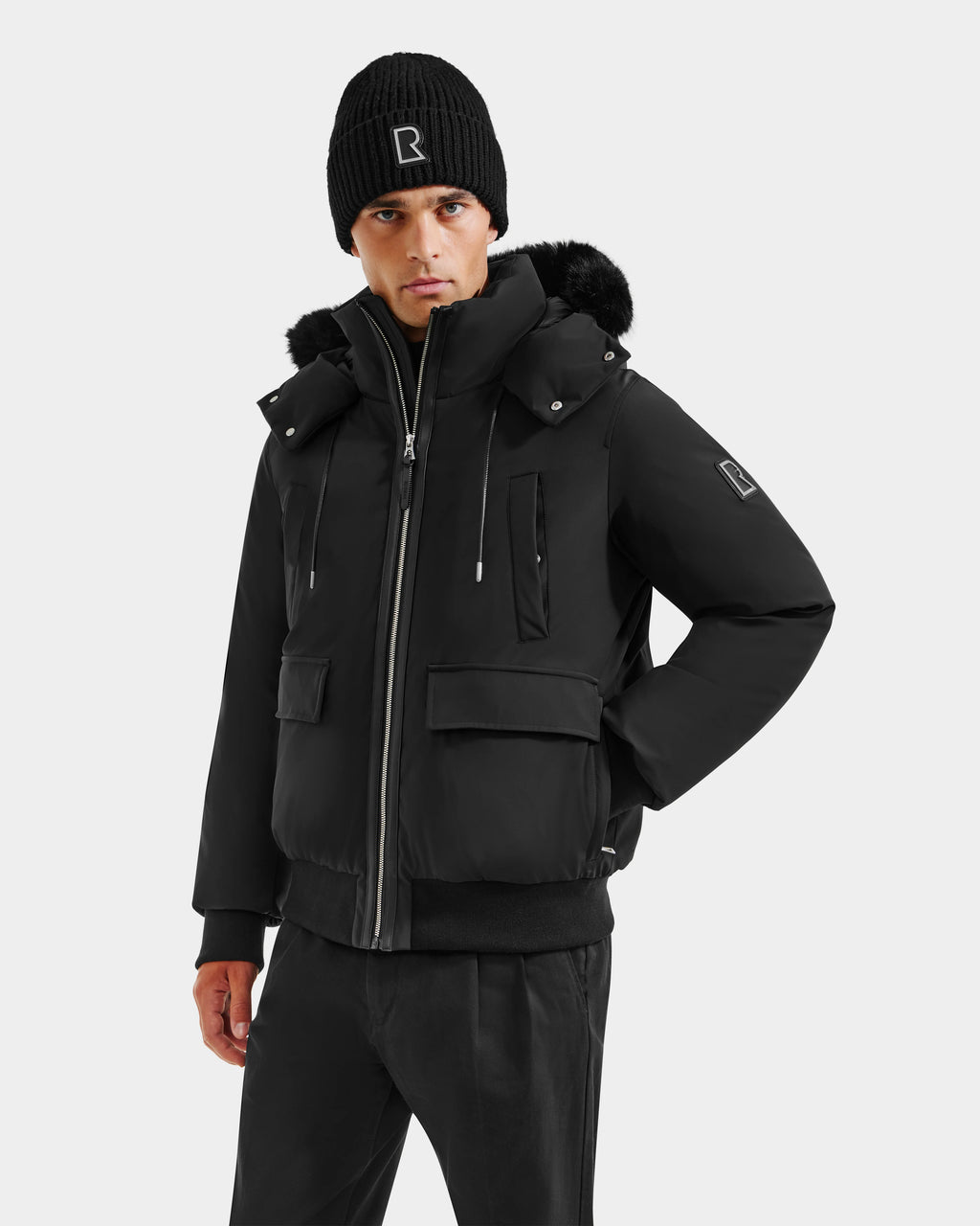 A man stands facing forward against a light background, wearing the Rudsak JEROME - 6125514 Black down bomber jacket with a fur-trimmed hood, paired with a black knit beanie and black pants, one hand in his pocket.