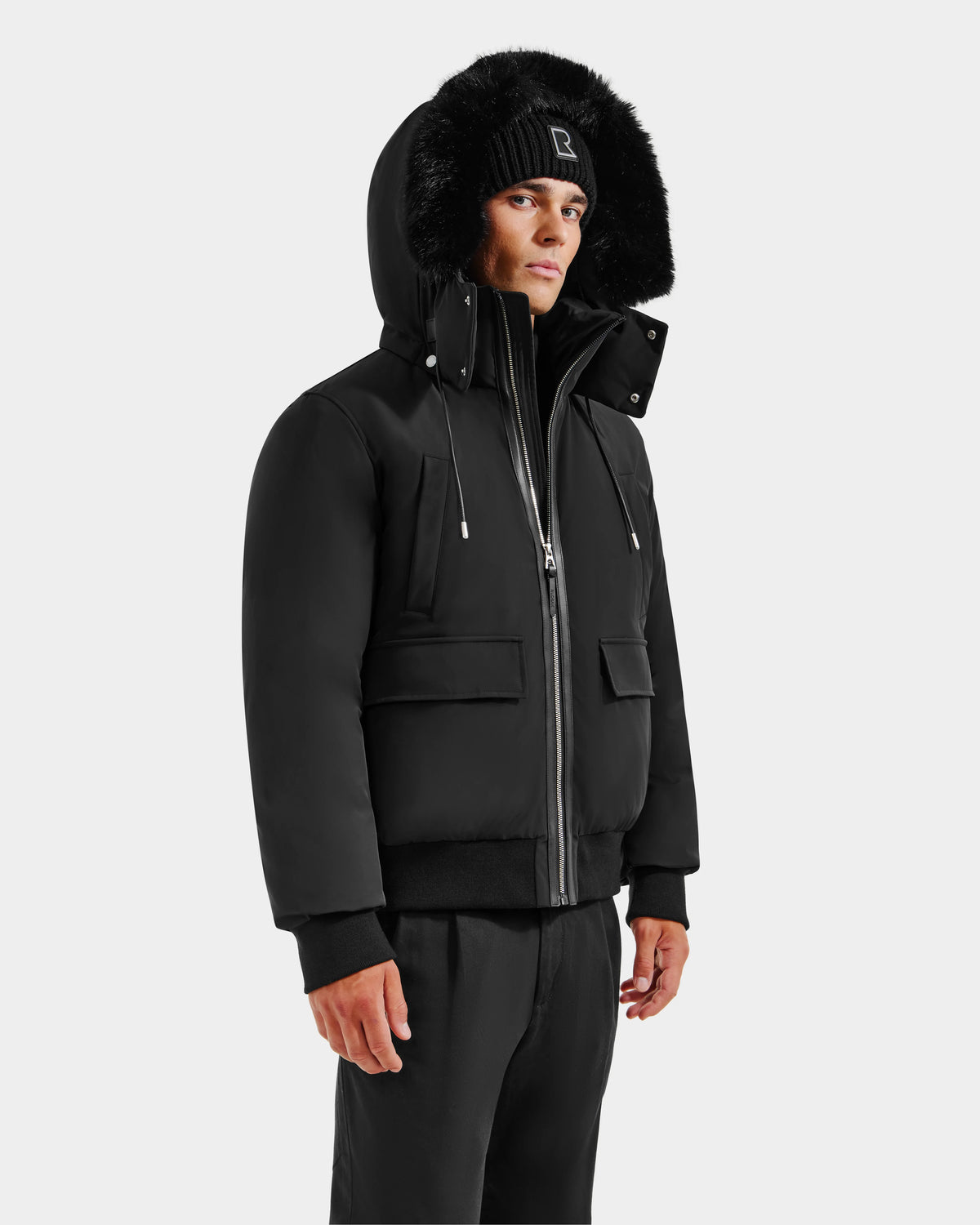 A person wearing the Rudsak JEROME - 6125514 Black men’s down bomber jacket with a fur-lined hood and a black knit beanie, standing against a plain white background.